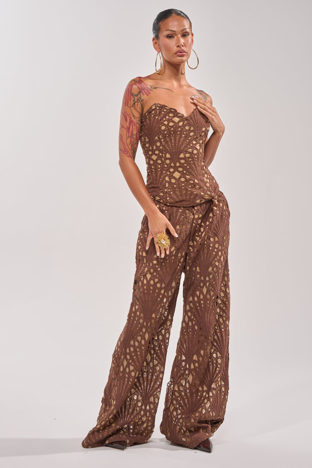 A person with large hoop earrings and tattoos models the CLAIRE EYELET PALAZZO PANT, featuring a strapless brown lace bodice and wide-leg fit, standing confidently against a plain light background. 5