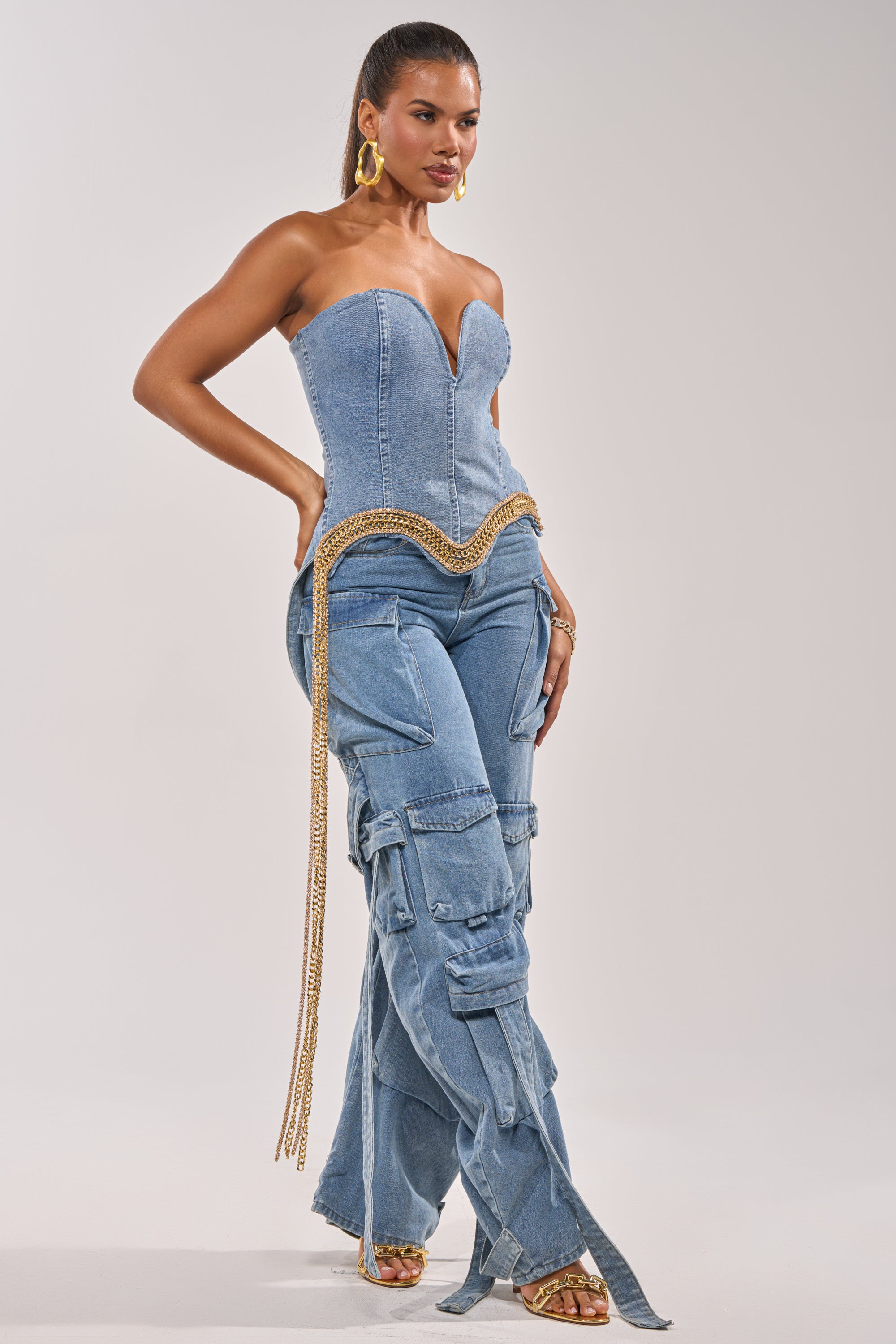 A woman poses confidently in the SHOW IT OFF DENIM CORSET, a strapless denim top with a sweetheart neckline, paired with wide-leg cargo denim pants. She completes her look with gold earrings, a chain belt, bracelets, and heeled sandals. 3