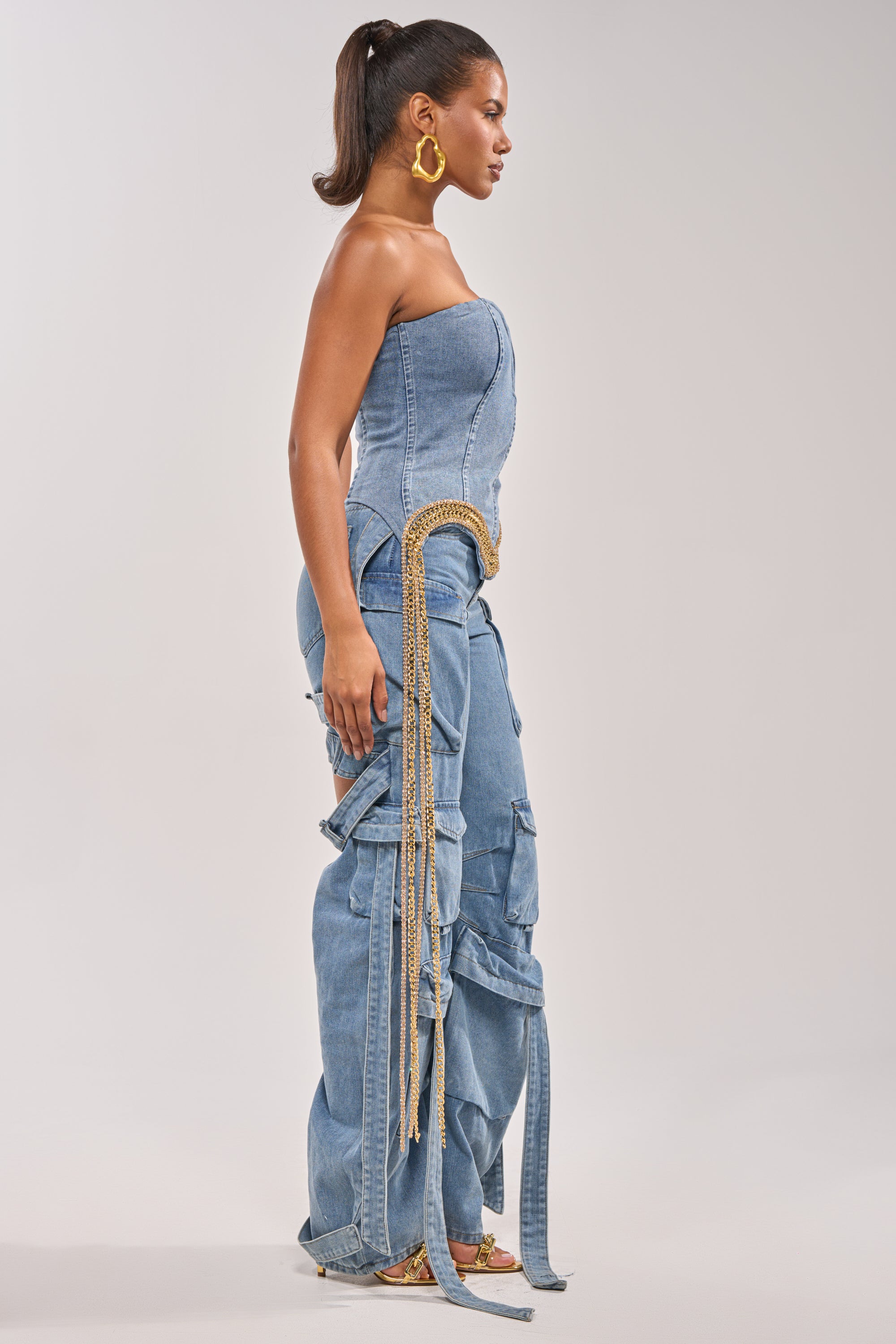A woman stands in profile wearing the SHOW IT OFF DENIM CORSET with a strapless sweetheart neckline, paired with matching wide-leg cargo pants, chain fringe trim, gold hoop earrings, and her hair in a high ponytail. 4