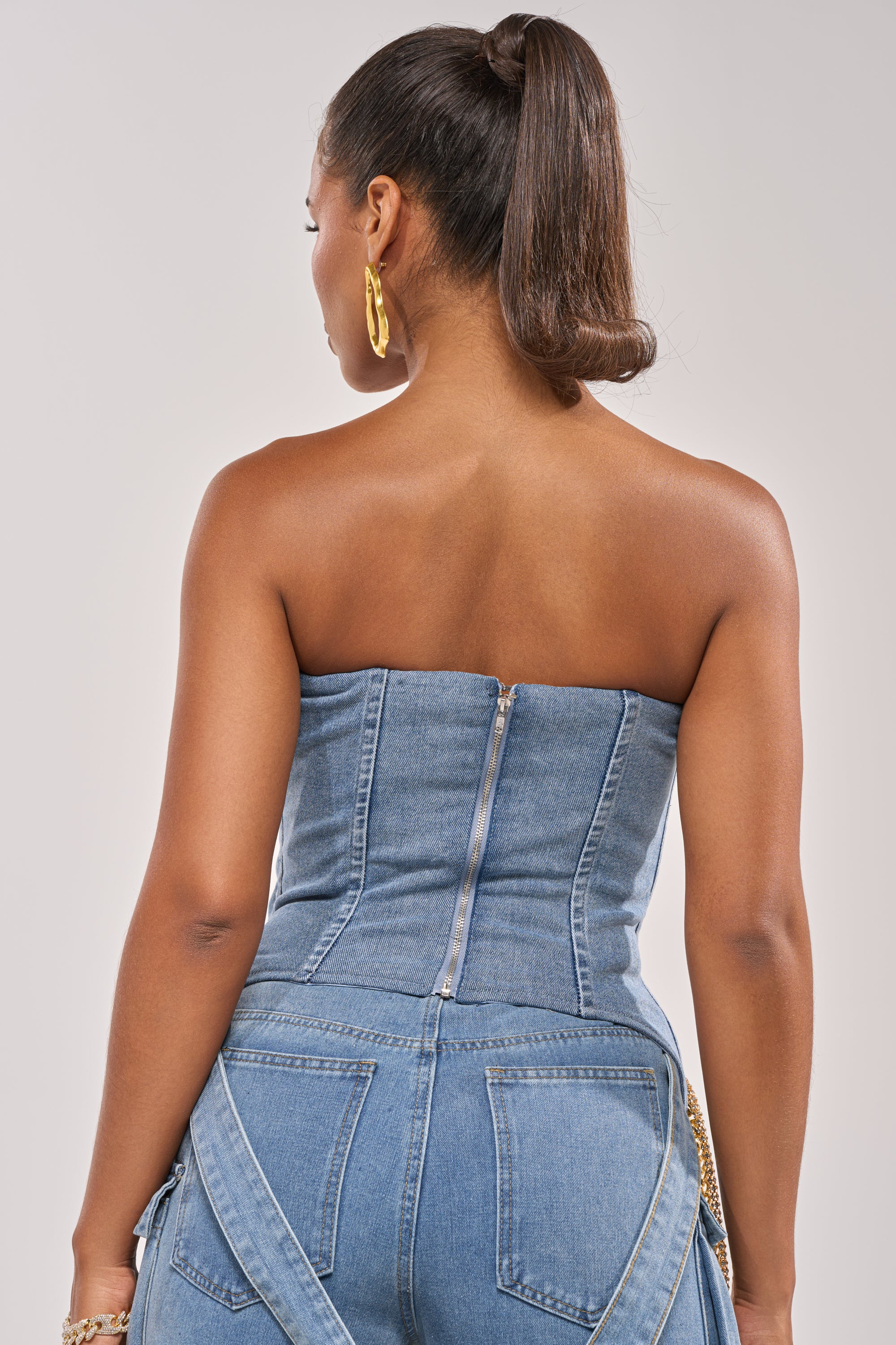 A woman with a high ponytail and large gold hoop earrings models the SHOW IT OFF DENIM CORSET and matching denim pants, shown from the back against a plain light background. 6