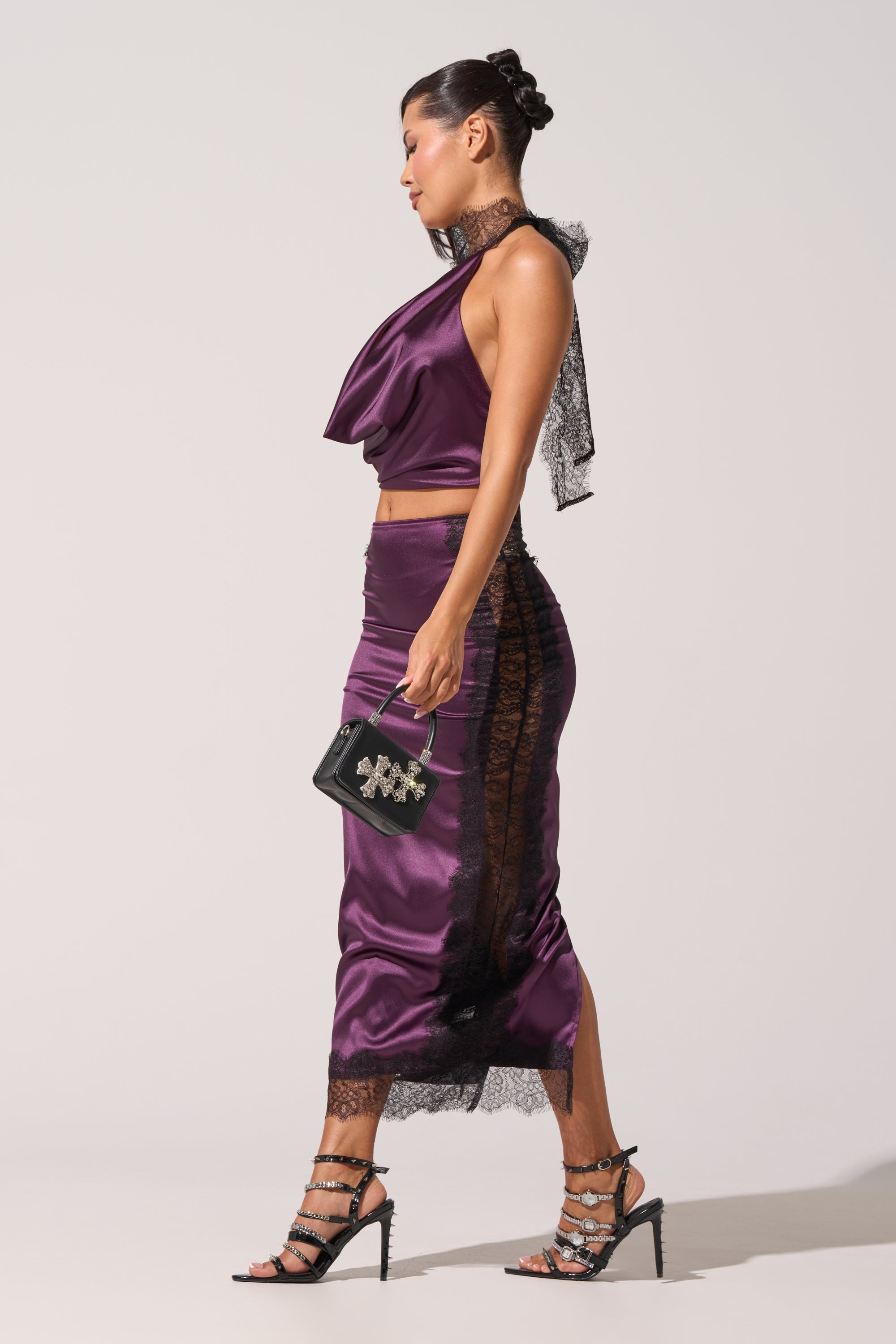 A woman in a shiny purple halter-neck top and the GOLDEN GIRL SATIN MAXI SKIRT with black lace trim walks in strappy high heels, carrying a small black handbag. Her hair is styled in an elegant updo. 1