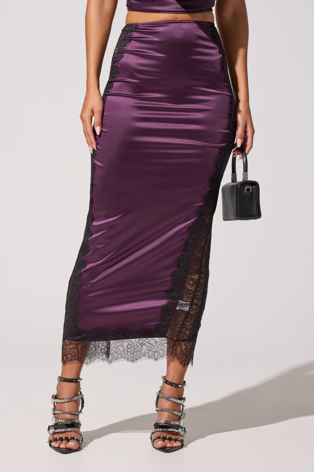 A woman models the GOLDEN GIRL SATIN MAXI SKIRT in shiny plum with black lace trim, paired with a small black handbag and strappy high-heeled sandals. The photo is cropped below her shoulders. 3