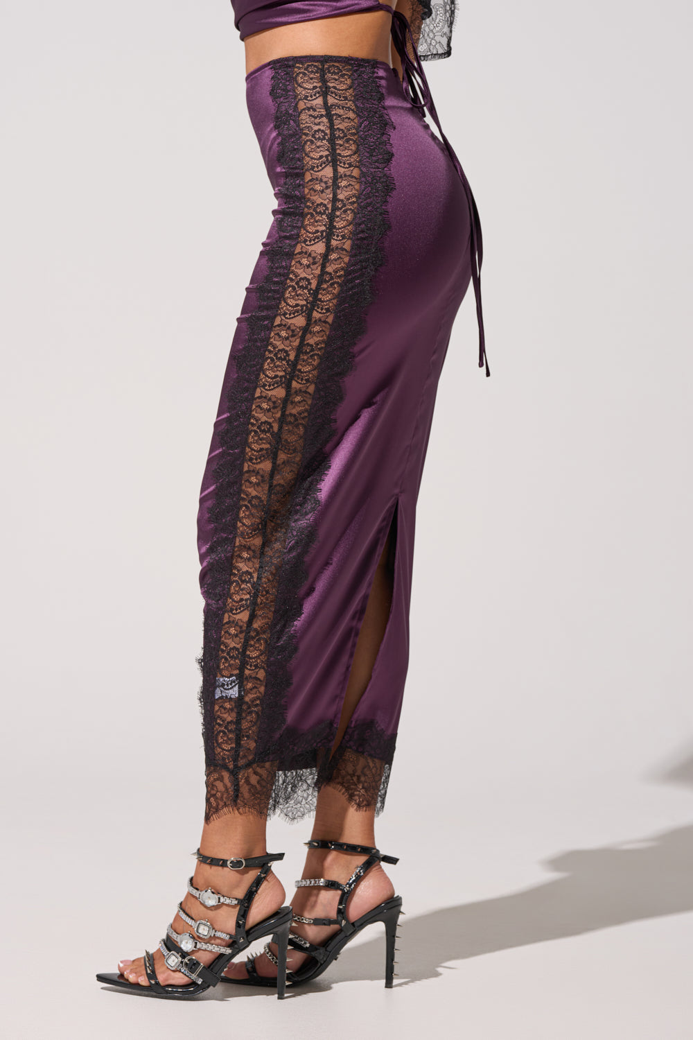 A person poses against a plain light background wearing the GOLDEN GIRL SATIN MAXI SKIRT in shiny purple satin with black lace trim, high-rise waist, side slit, and black rhinestone-strap high-heeled sandals. 4