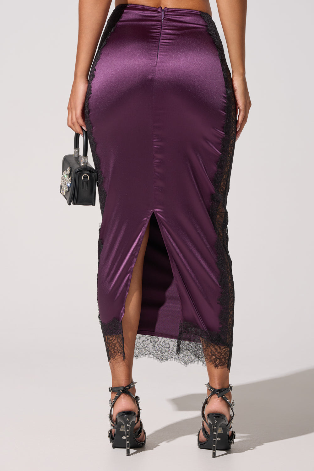 A person wearing the GOLDEN GIRL SATIN MAXI SKIRT—fitted, shiny purple with black lace trim and a back slit—paired with a small black handbag and decorative black high-heeled sandals. 5
