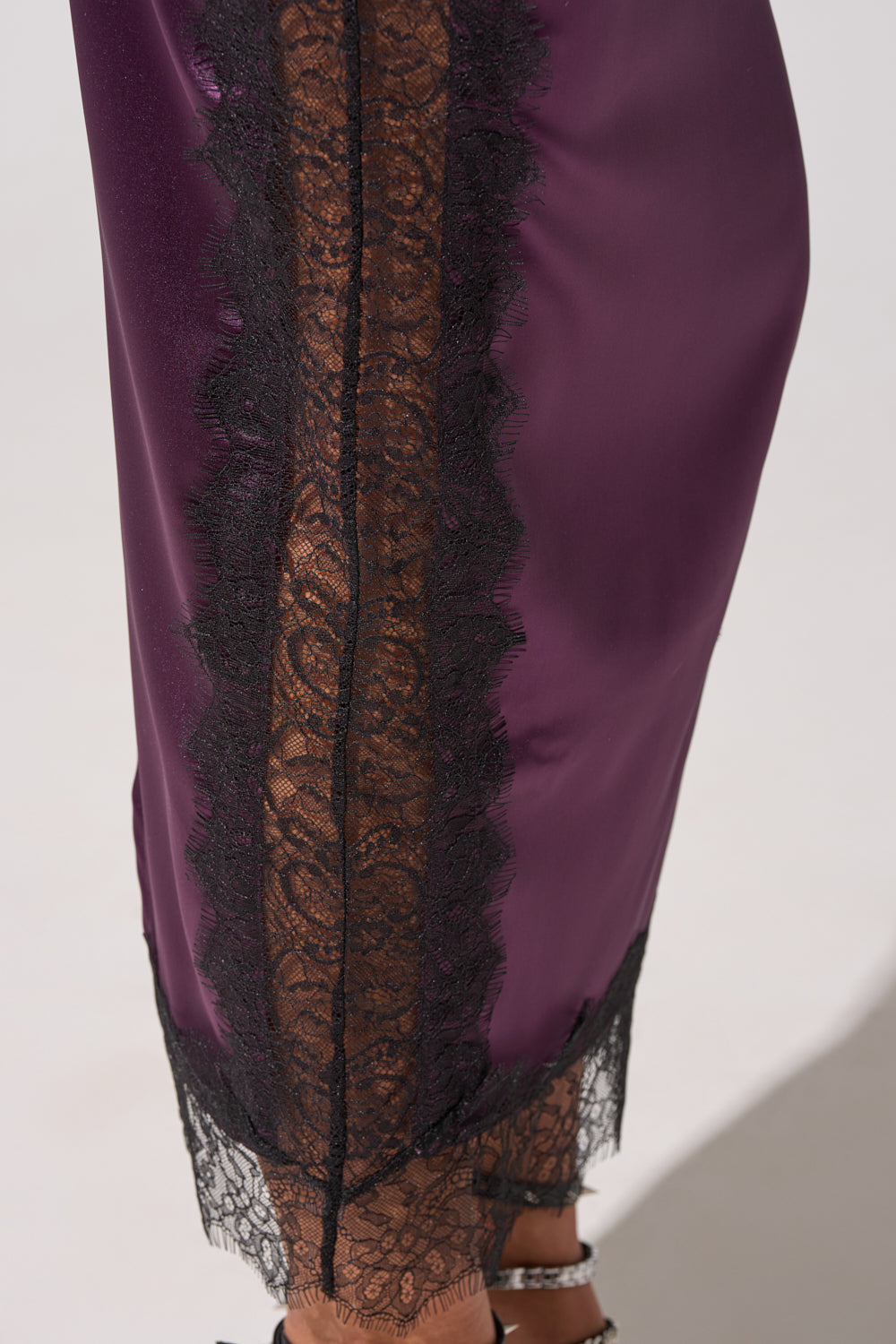 A close-up of the GOLDEN GIRL SATIN MAXI SKIRT, a purple satin maxi with a high-rise waist, vertical black lace trim, and black lace at the hem, styled with strappy heels. 7