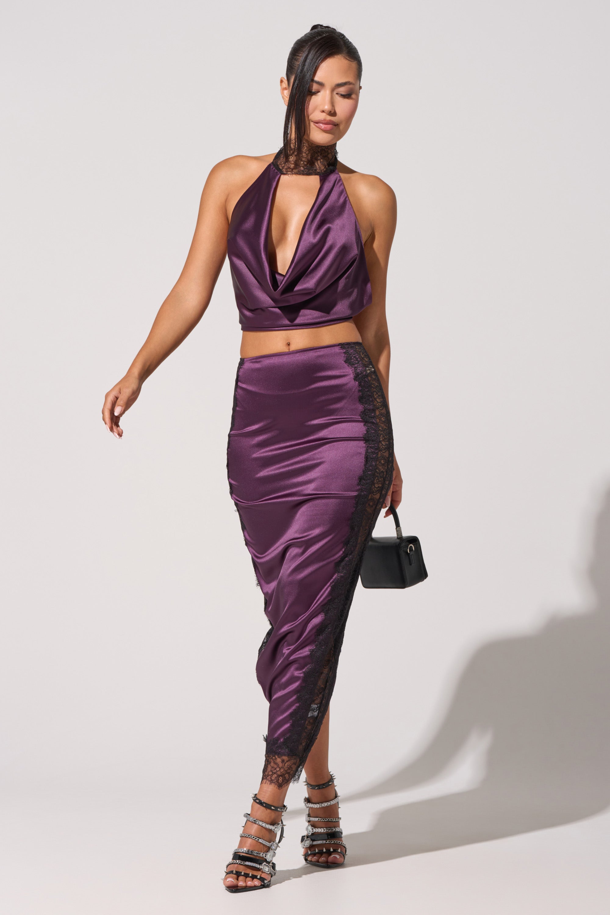A woman poses in the GOLDEN GIRL SATIN BLOUSE, a shiny plum halter-neck satin top paired with a matching high-waisted skirt trimmed in black lace, accessorized with a small black handbag and strappy high heels against a plain light background. 1