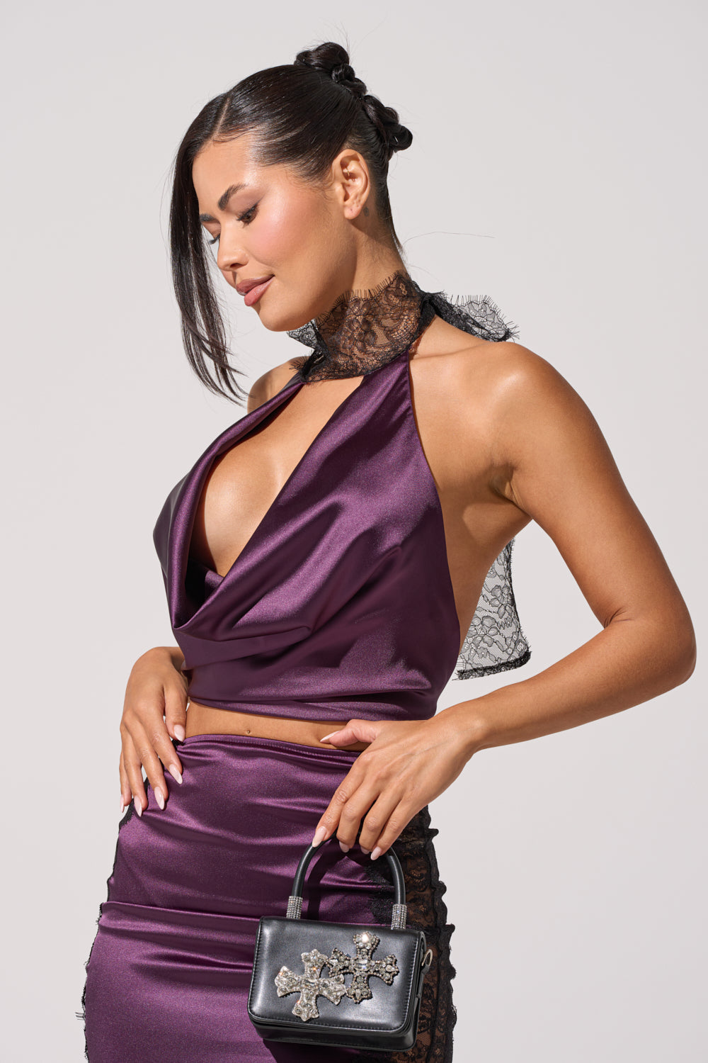 A woman wears the GOLDEN GIRL SATIN BLOUSE, a shiny purple halter top with an open-back design, paired with a matching lace-detailed skirt. She carries a jeweled black handbag, has her hair up, and sports neutral makeup. 2