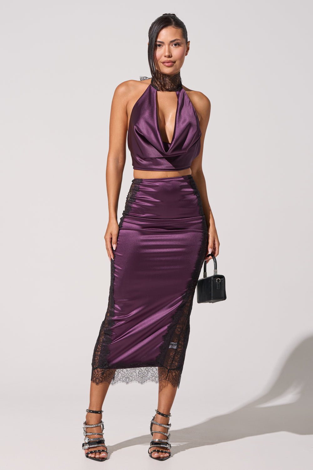 A woman wearing the GOLDEN GIRL SATIN BLOUSE, a shiny purple open-back top, with a matching high-waisted midi skirt featuring black lace trim, holds a small black purse and wears strappy heels against a plain light background. 5