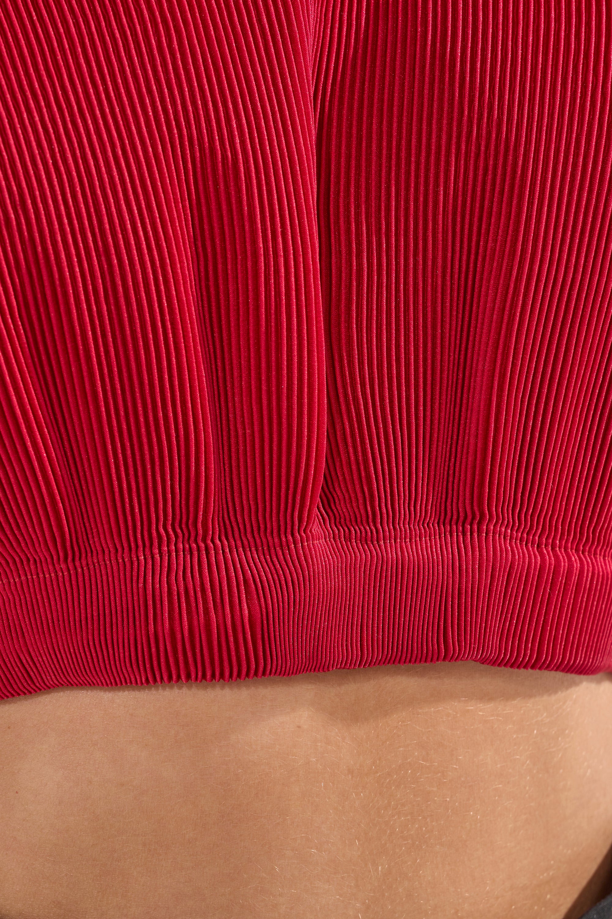 Close-up of the ALL ABOUT THE DRAMA TOP, a red pleated crop top with bold ribbed texture and visible folds, sitting above exposed skin at the waist for a striking fashion statement. 6