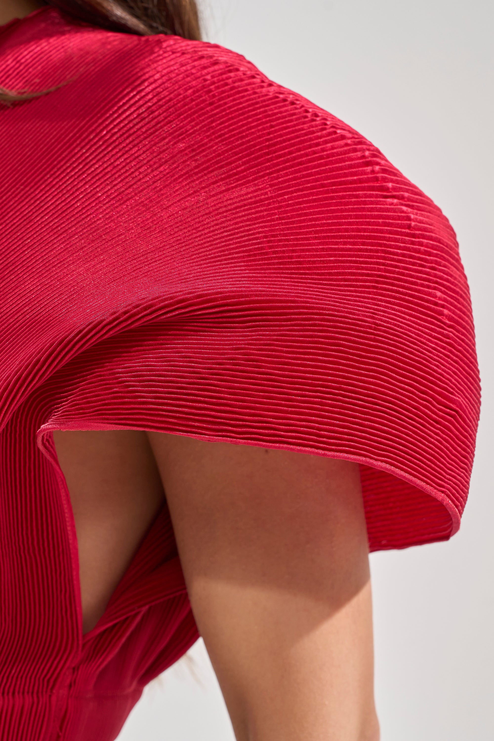 A close-up of a person wearing the ALL ABOUT THE DRAMA TOP—a sculptural, pleated red crop top with batwing sleeves curving outward, revealing part of their shoulder and upper arm for an undeniable statement look. 7