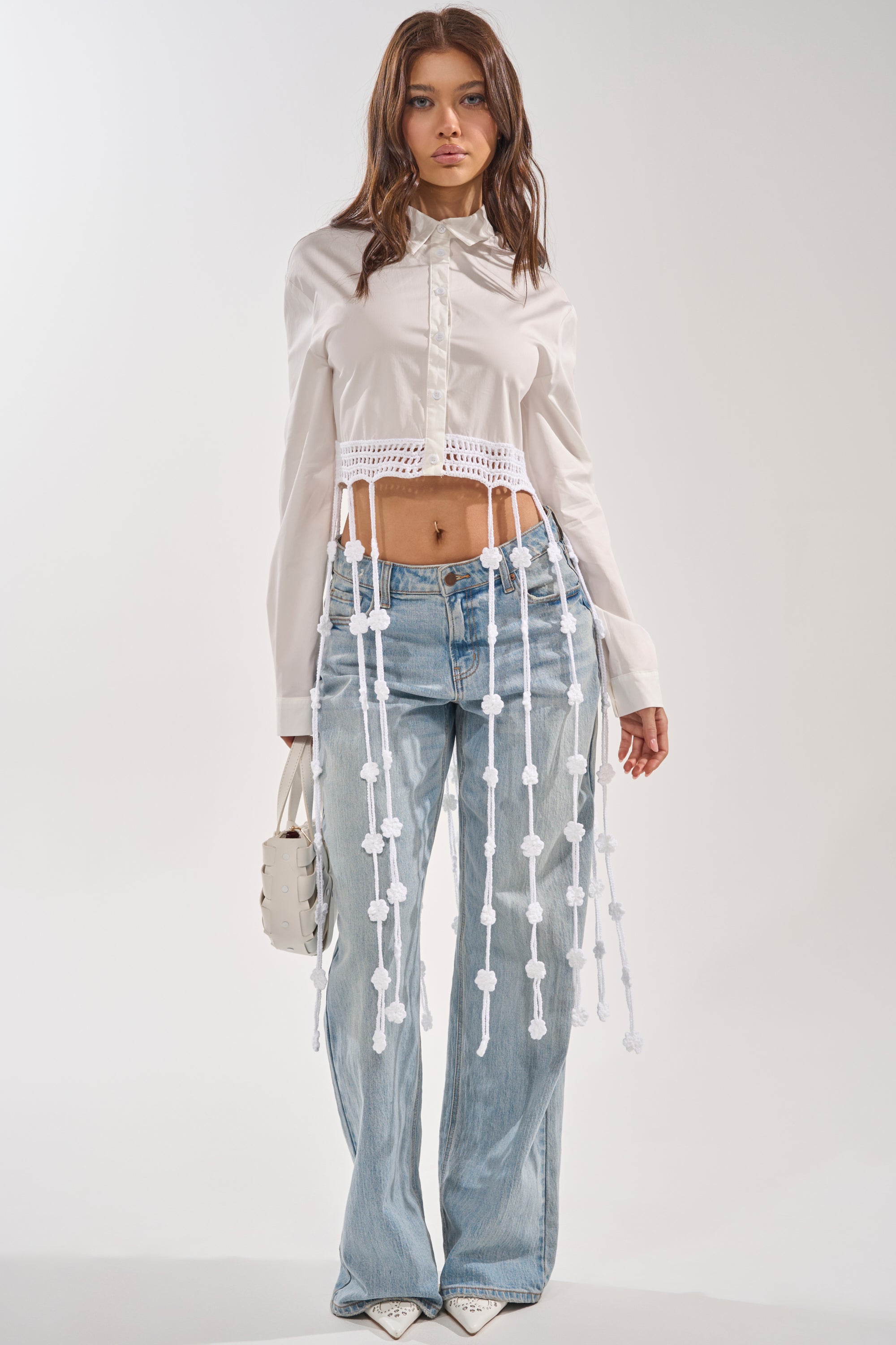 A woman faces forward in a PETAL DRIFT CROCHET DETAIL BLOUSE, light blue wide-leg jeans, white pointed shoes, and holding a small white handbag against a plain light background. 0