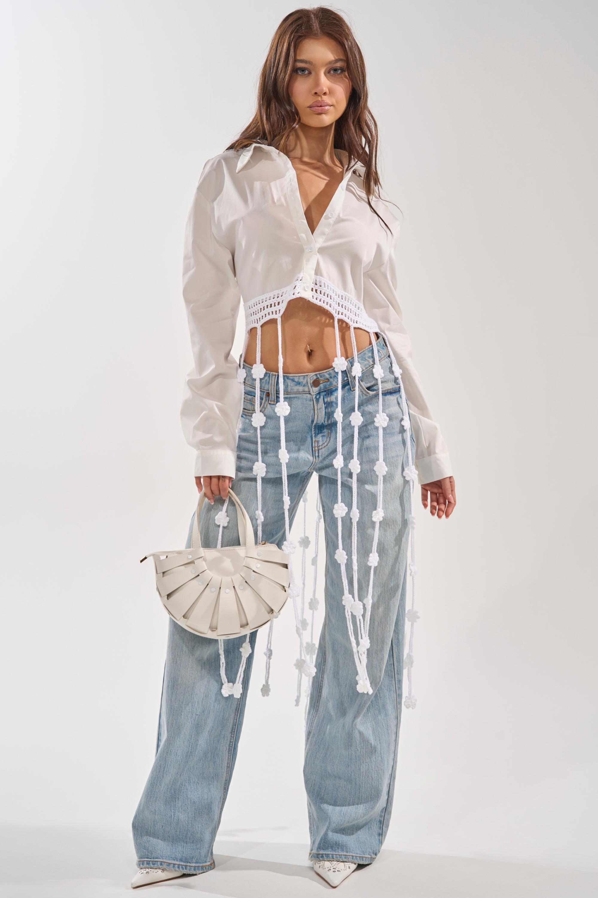 A woman models the PETAL DRIFT CROCHET DETAIL BLOUSE—a white button down shirt with long crochet fringe—styled with light blue wide-leg jeans, pointed white shoes, and a round white bag for an effortless festival look. 1