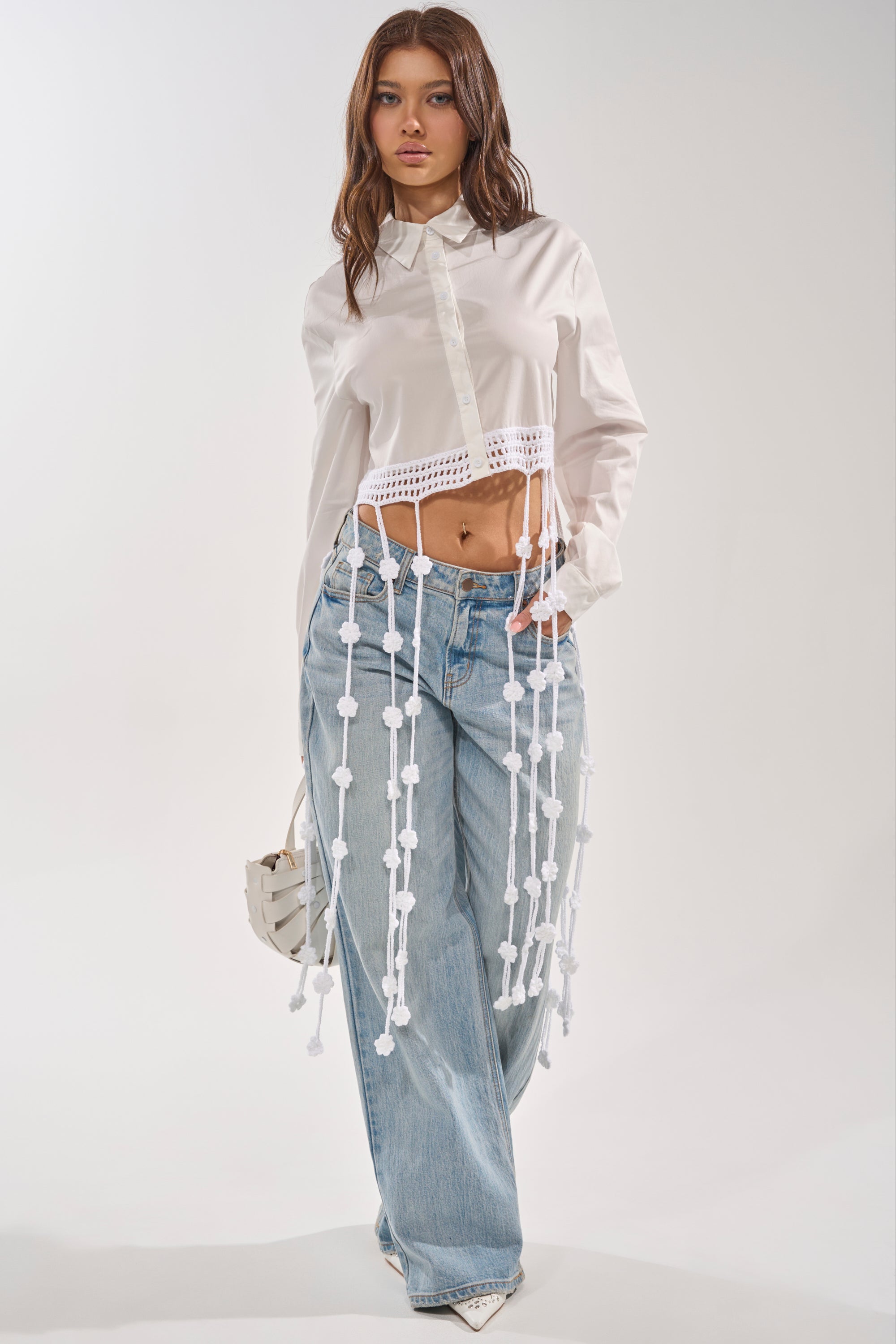 She wears the PETAL DRIFT CROCHET DETAIL BLOUSE, a white button-down with fringe, long decorative strings and beads, styled with light blue wide-leg jeans, white pointed-toe shoes, and a small white handbag—perfect festival wear. 2