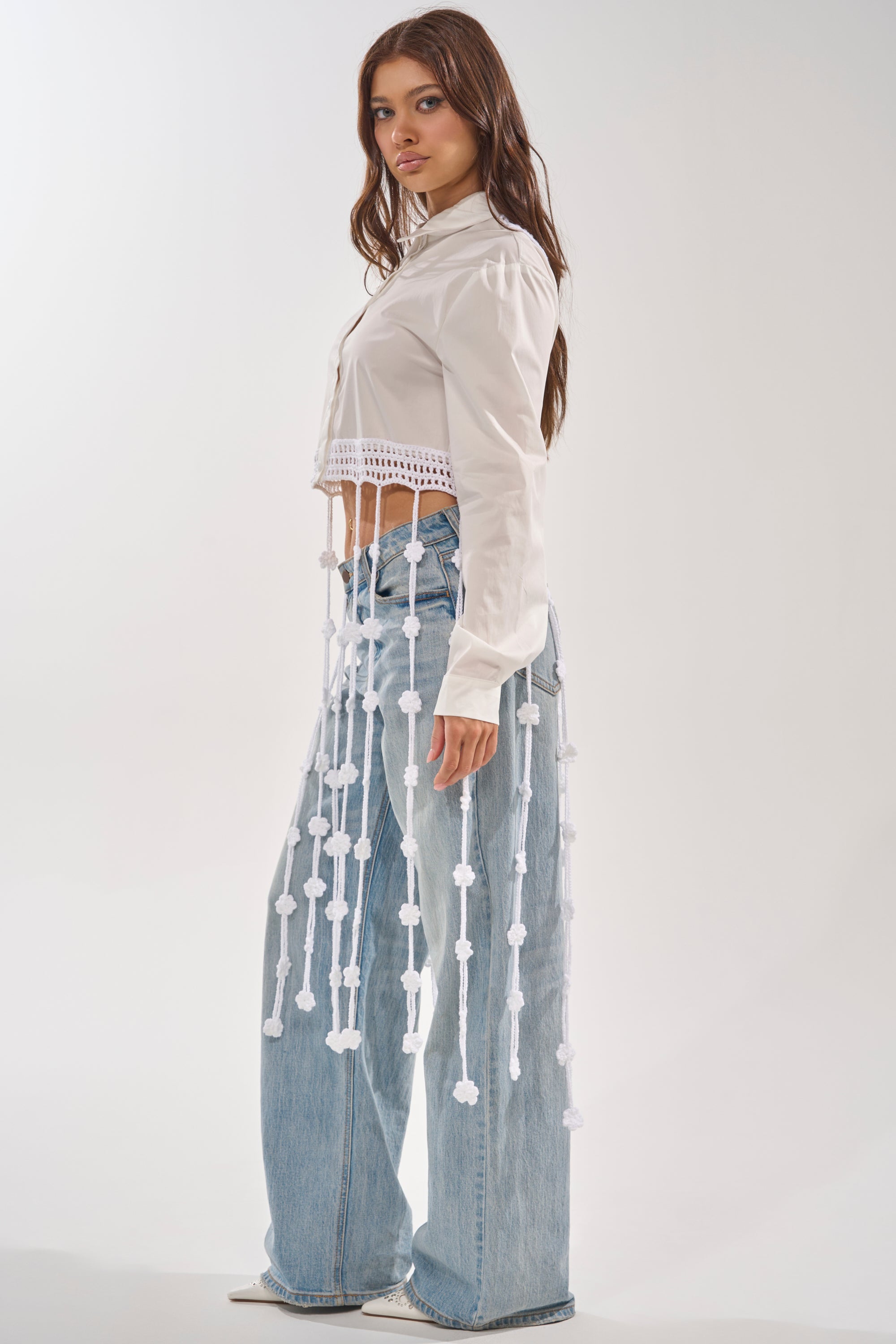 A woman stands sideways against a white background wearing the PETAL DRIFT CROCHET DETAIL BLOUSE, a white cropped button-down with tasseled trim, paired with high-waisted, wide-leg light blue jeans—an ideal festival look. 3
