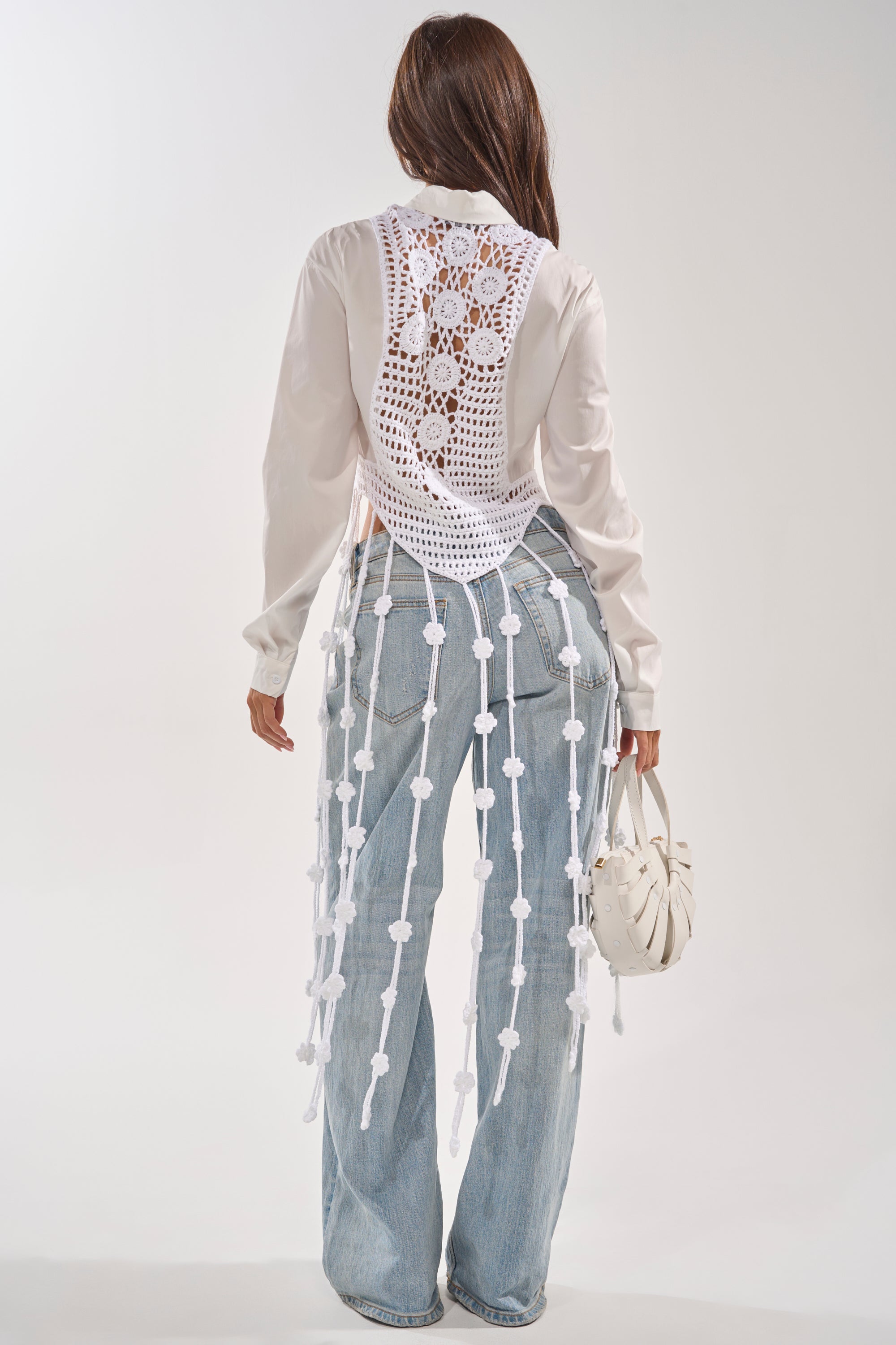 A woman with long brown hair faces away, wearing the PETAL DRIFT CROCHET DETAIL BLOUSE as a festival top over a white blouse and light blue wide-legged jeans, paired with a small white handbag. 4
