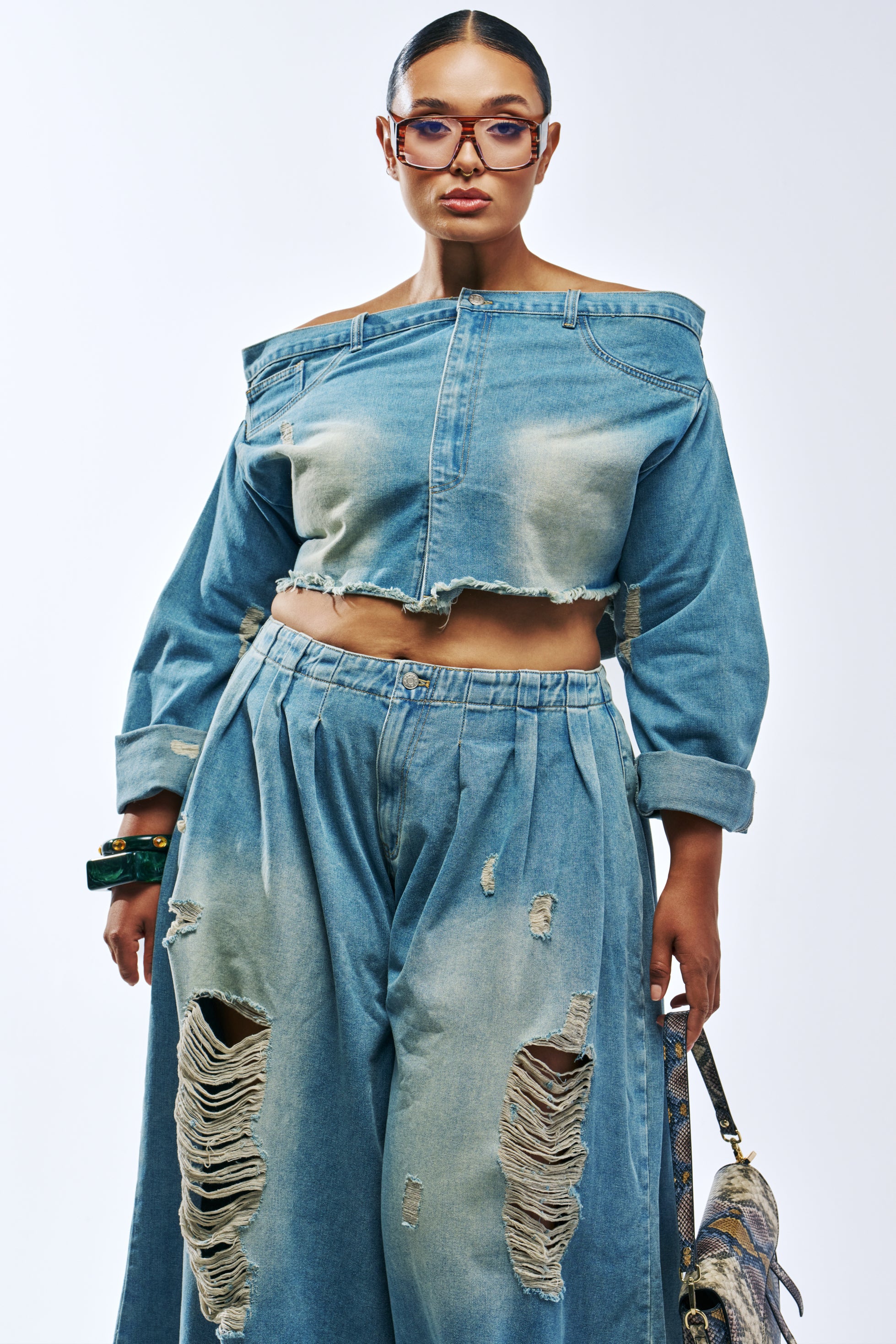 A model wears the CAMILLE DENIM BLOUSE IN MEDIUM BLUE DENIM, an off-the-shoulder, cropped, medium-wash distressed denim top with raw edges.