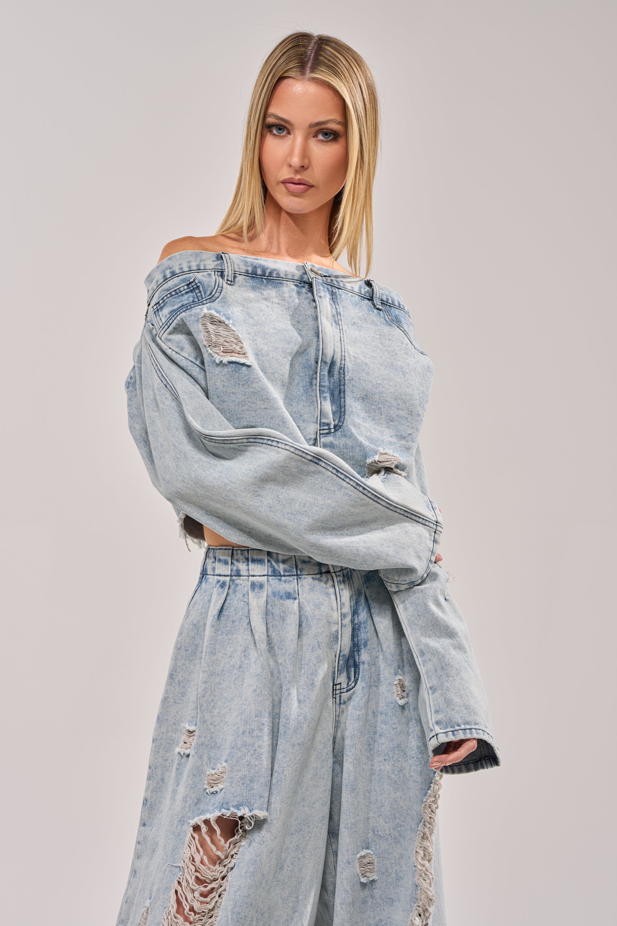 A woman with straight blonde hair models the CAMILLE DENIM BLOUSE, a loose-fitting, off-the-shoulder top, paired with ripped denim pants against a plain light background. 0
