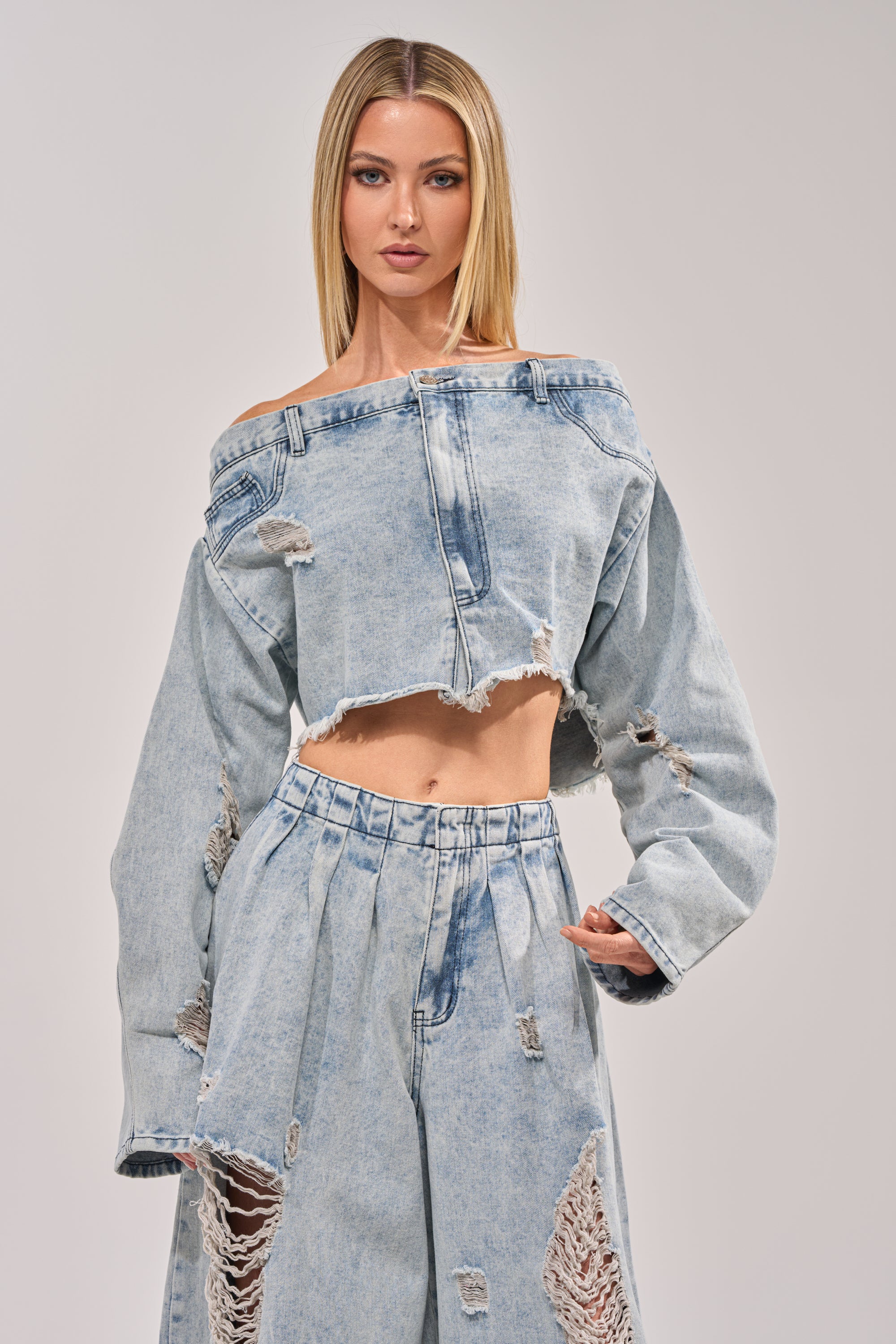 A woman with straight blonde hair and a neutral expression wears the CAMILLE DENIM BLOUSE, featuring an off-the-shoulder, distressed, cropped raw hem design, paired with matching high-waisted wide-leg jeans. 1