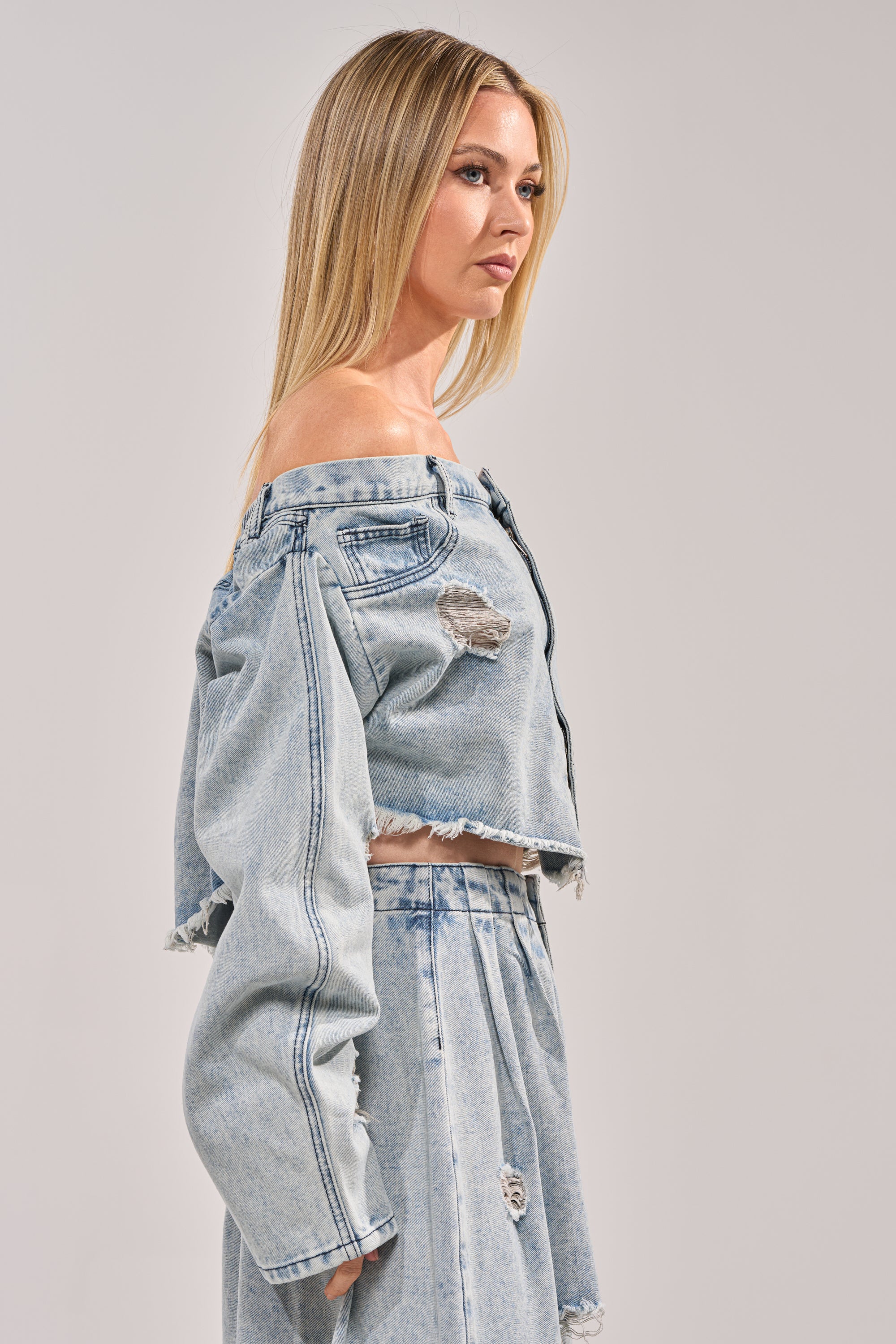 A woman with long blonde hair stands in profile against a plain background, wearing the CAMILLE DENIM BLOUSE—an off-the-shoulder, distressed denim top paired with matching high-waisted pants featuring a cropped raw hem. 2