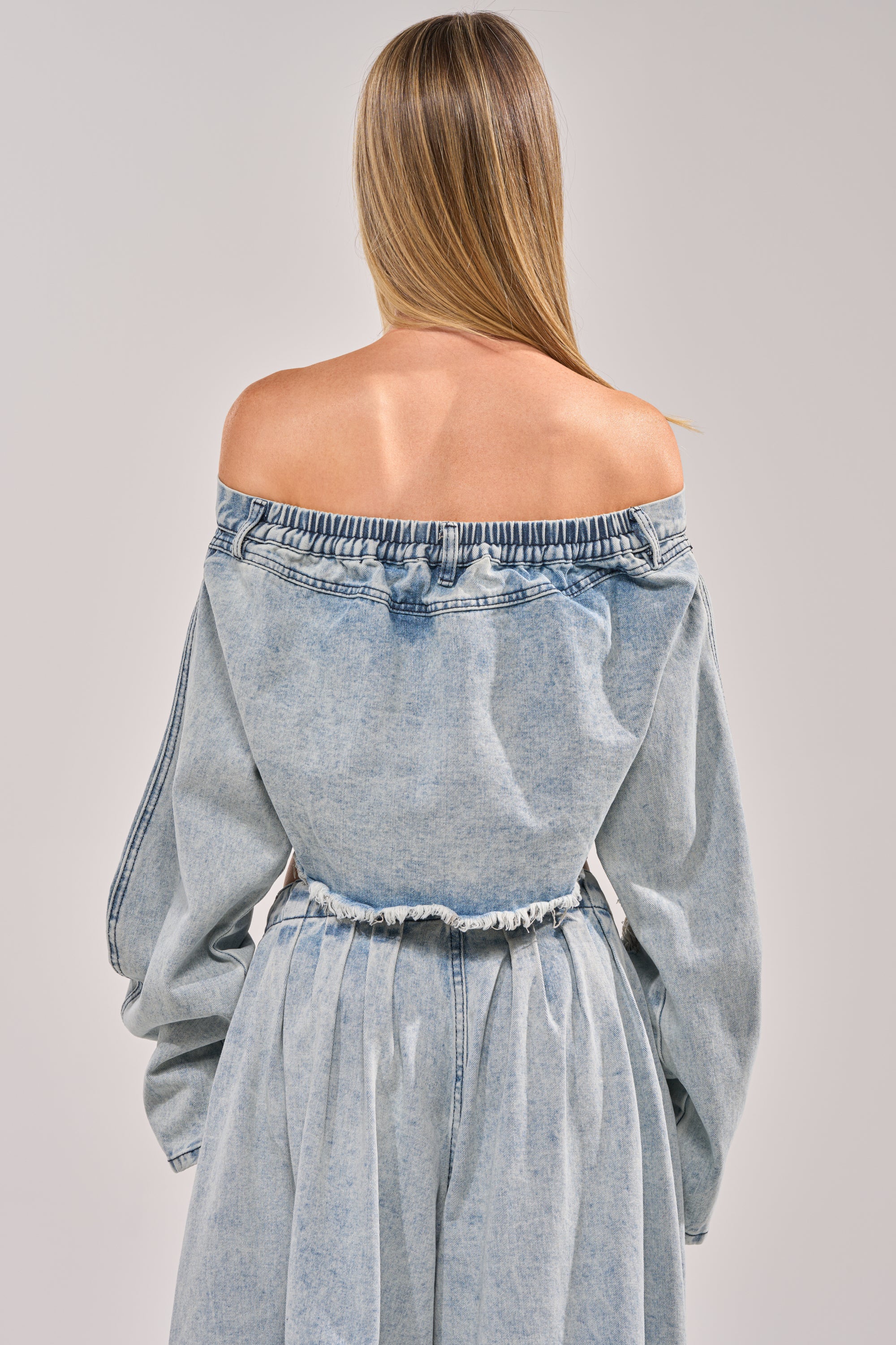 A woman with straight blonde hair is seen from behind, wearing the CAMILLE DENIM BLOUSE—an off-the-shoulder, light blue top with long sleeves, elastic neckline, gathered waist, and a subtle cropped raw hem against a plain light background. 3
