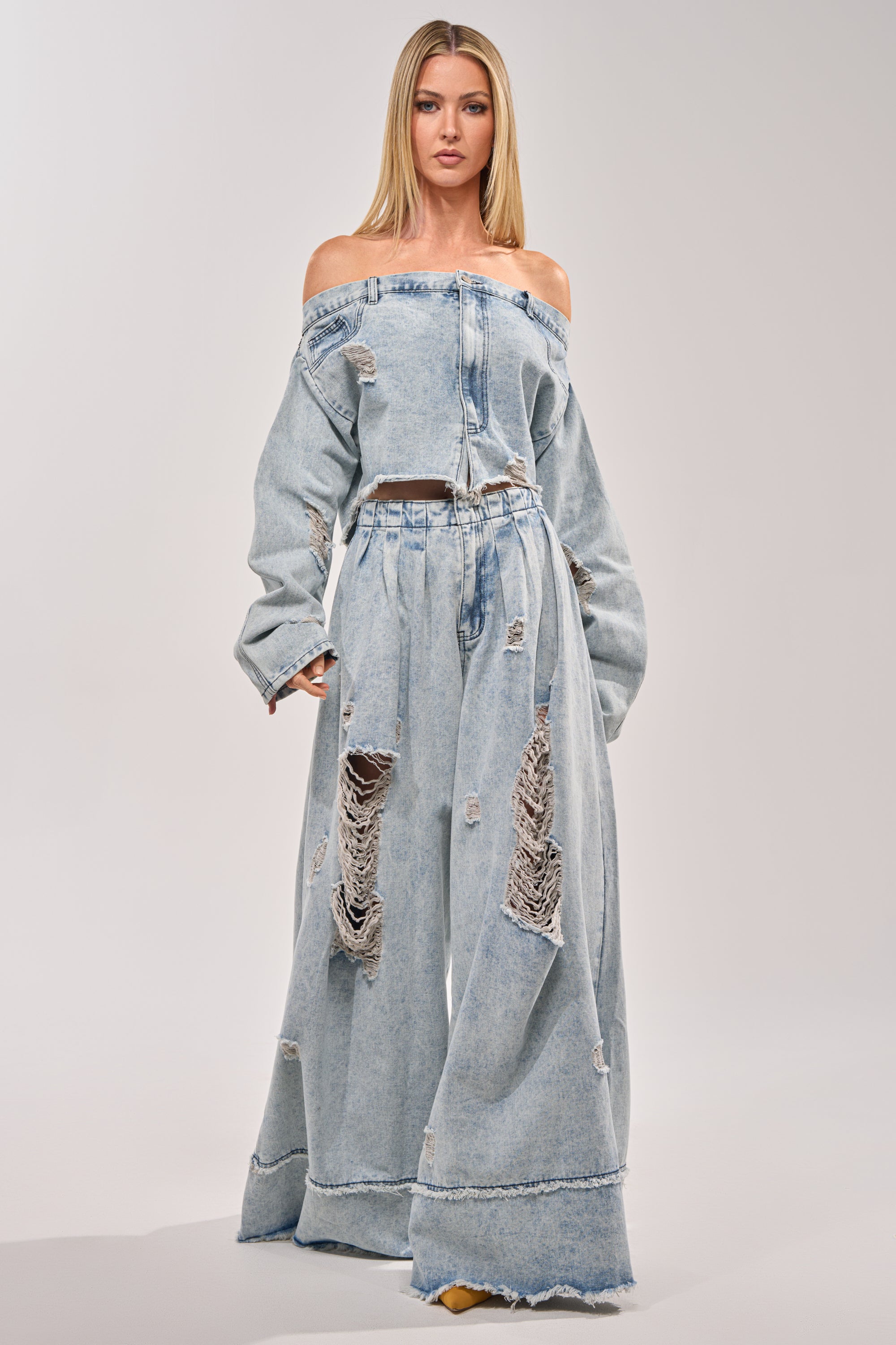 Wearing the CAMILLE DENIM BLOUSE, a woman pairs this off-shoulder, long-sleeve distressed top with wide-leg, high-waisted ripped jeans with a cropped raw hem as she stands before a plain light background. 5