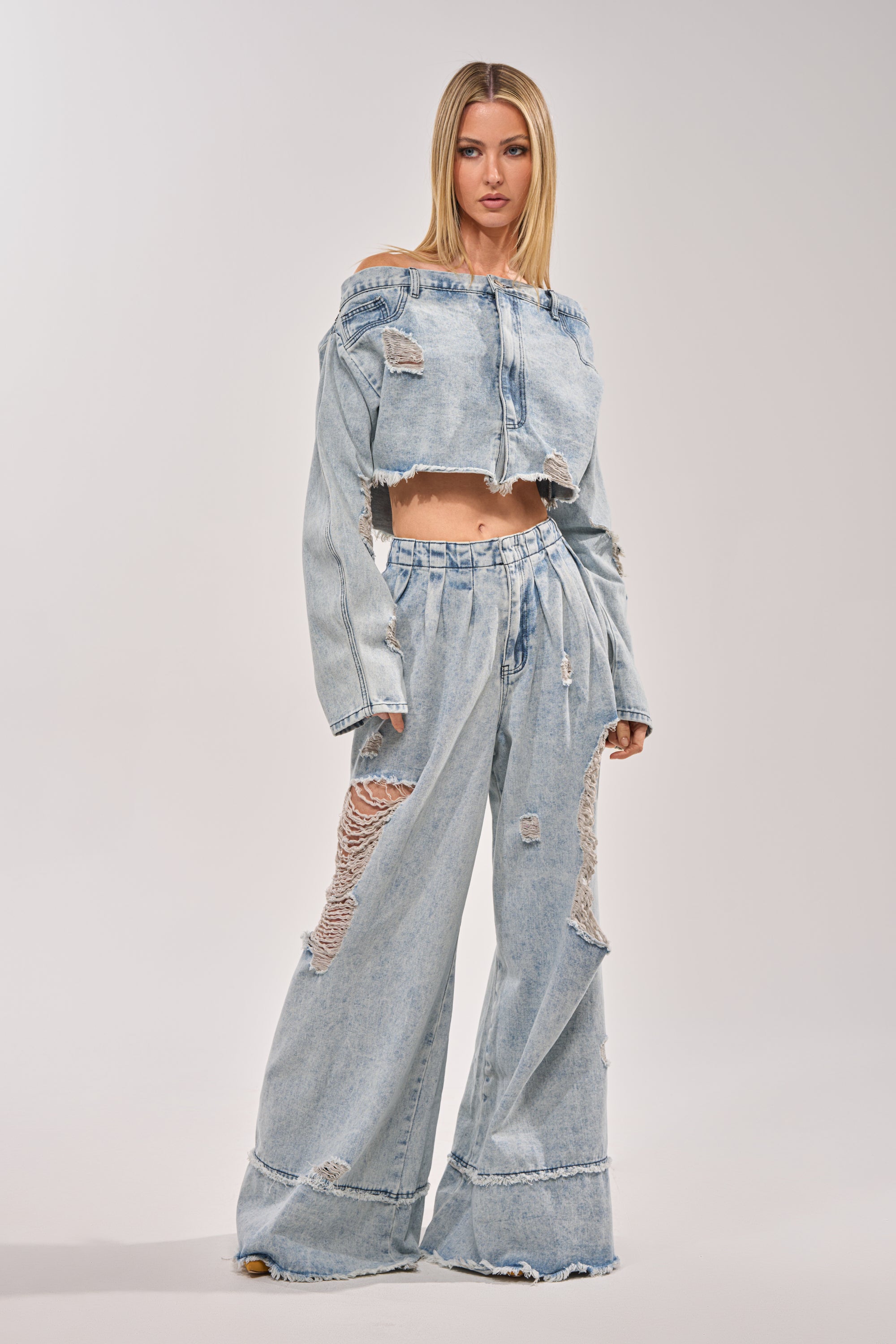 A woman wears the CAMILLE DENIM BLOUSE, a light blue, off-the-shoulder top with a cropped raw hem, paired with matching wide-leg pants featuring large rips and frayed edges, standing against a plain background. 6