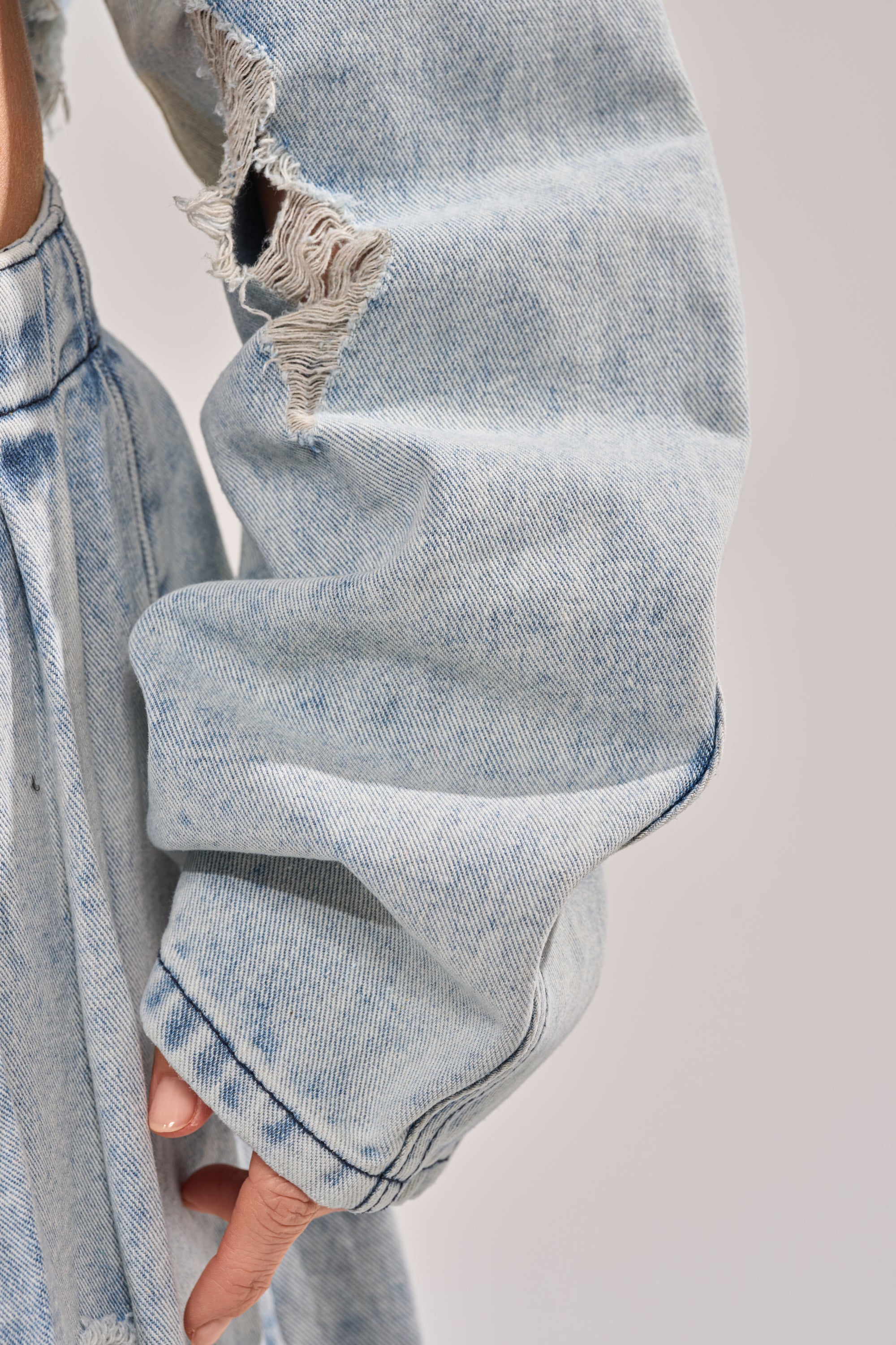 A close-up of a person wearing the CAMILLE DENIM BLOUSE, showcasing its ripped sleeve and frayed, faded blue denim for an edgy, distressed style. 7