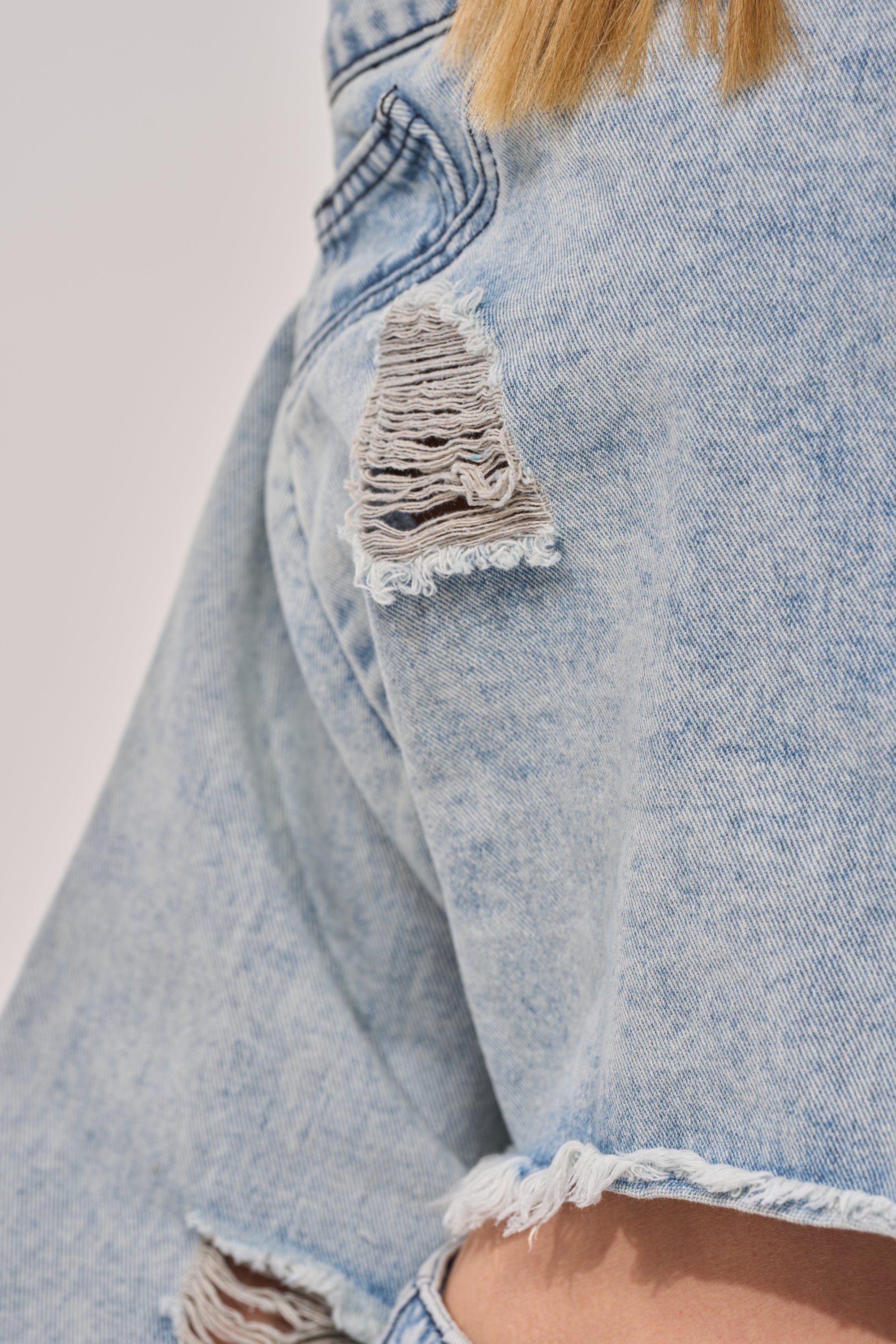 Close-up of a person wearing the CAMILLE DENIM BLOUSE, featuring light blue denim with frayed details and a cropped raw hem for an edgy look. Some light brown hair is visible near the top of the image. 8