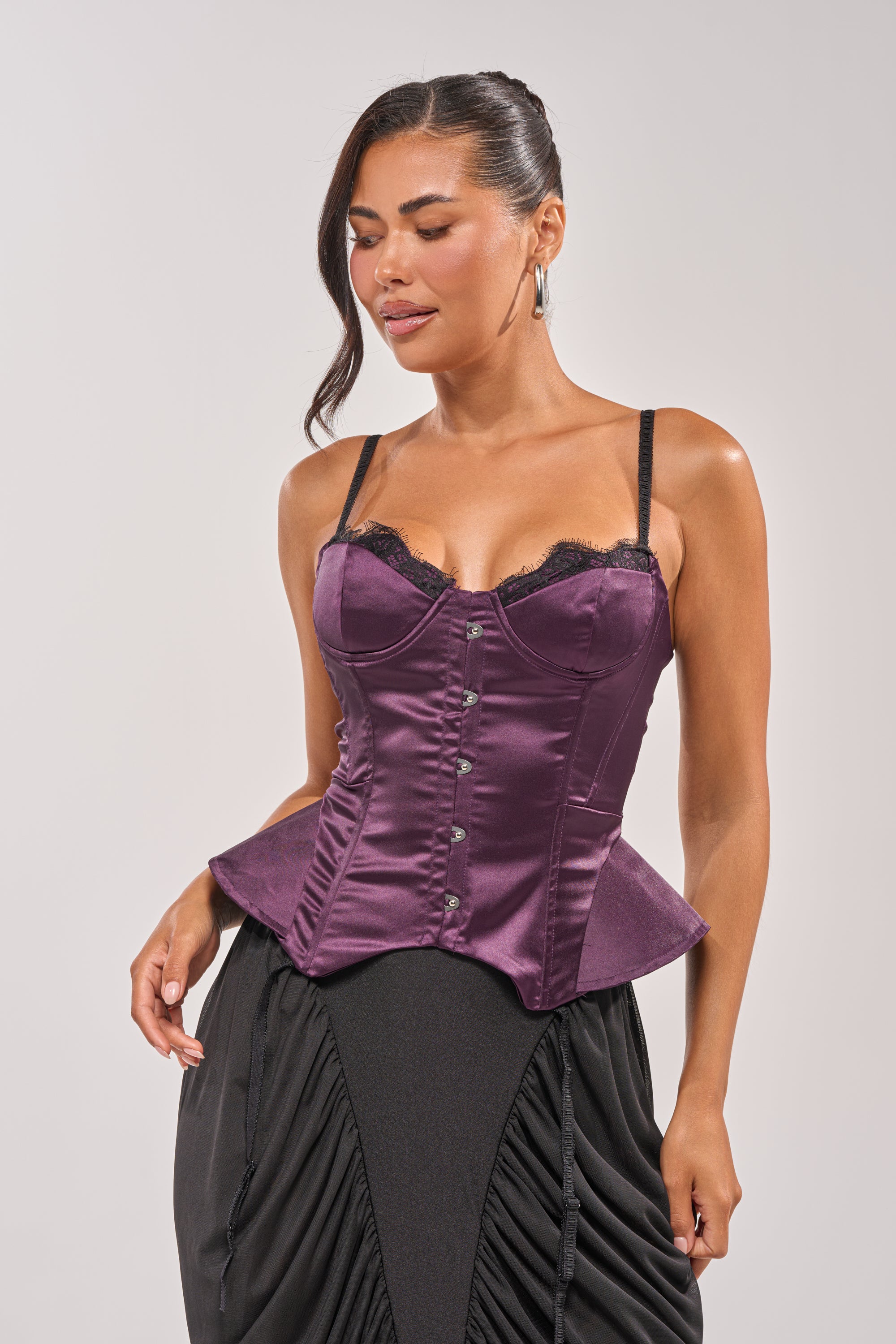 She wears the FLY ME OUT SATIN CORSET, a shiny purple top with black lace trim and thin straps, styled with a black skirt. Her long dark hair is swept to one side as she stands against a light background. 1