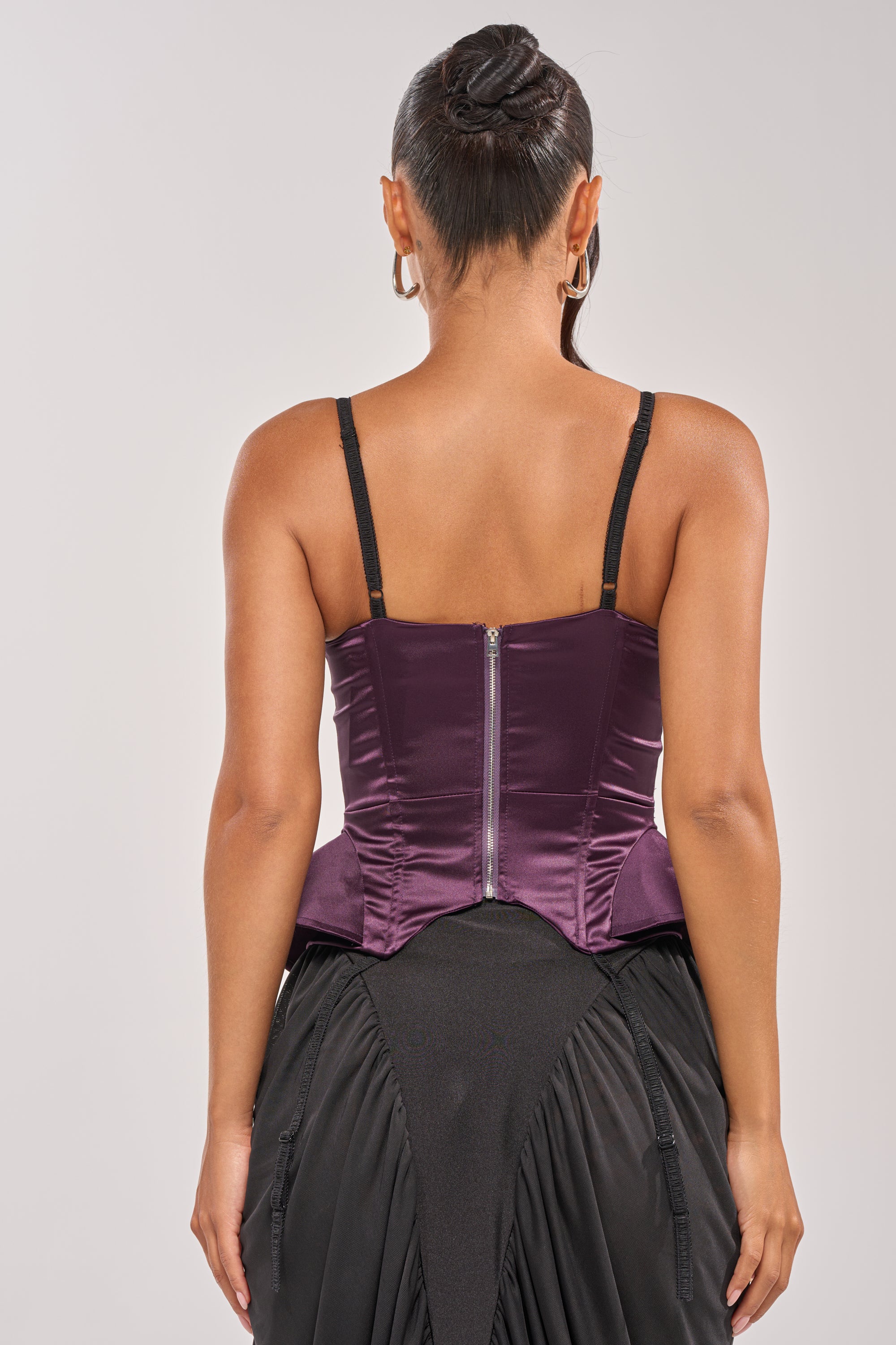 A woman is seen from the back in the FLY ME OUT SATIN CORSET, a shiny purple peplum top with black straps and exposed zipper, paired with a black skirt. She sports a sleek updo and hoop earrings. 3
