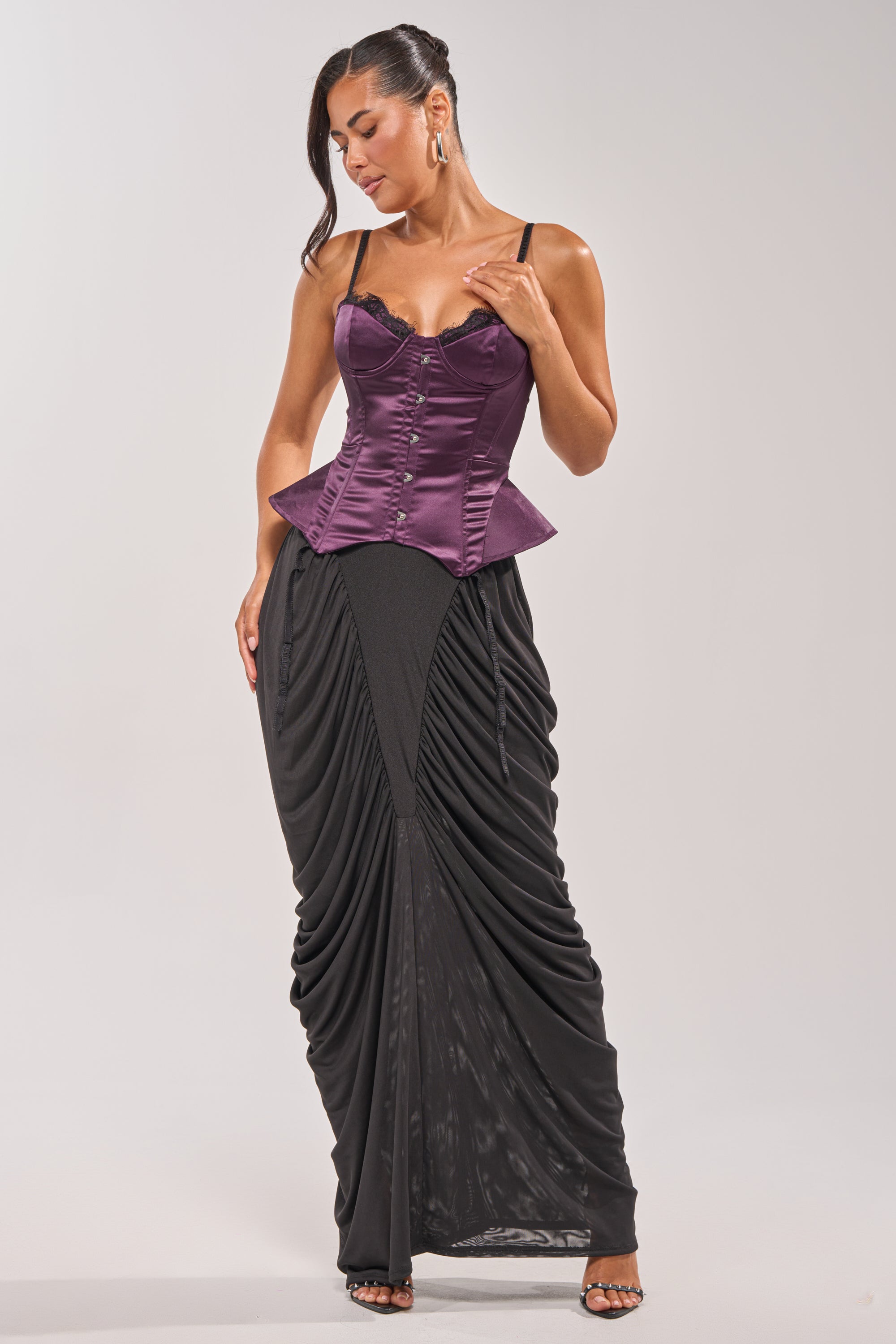A woman poses against a plain light background wearing the FLY ME OUT SATIN CORSET in purple with thin straps, styled with a long black draped skirt and black open-toe heels. 4