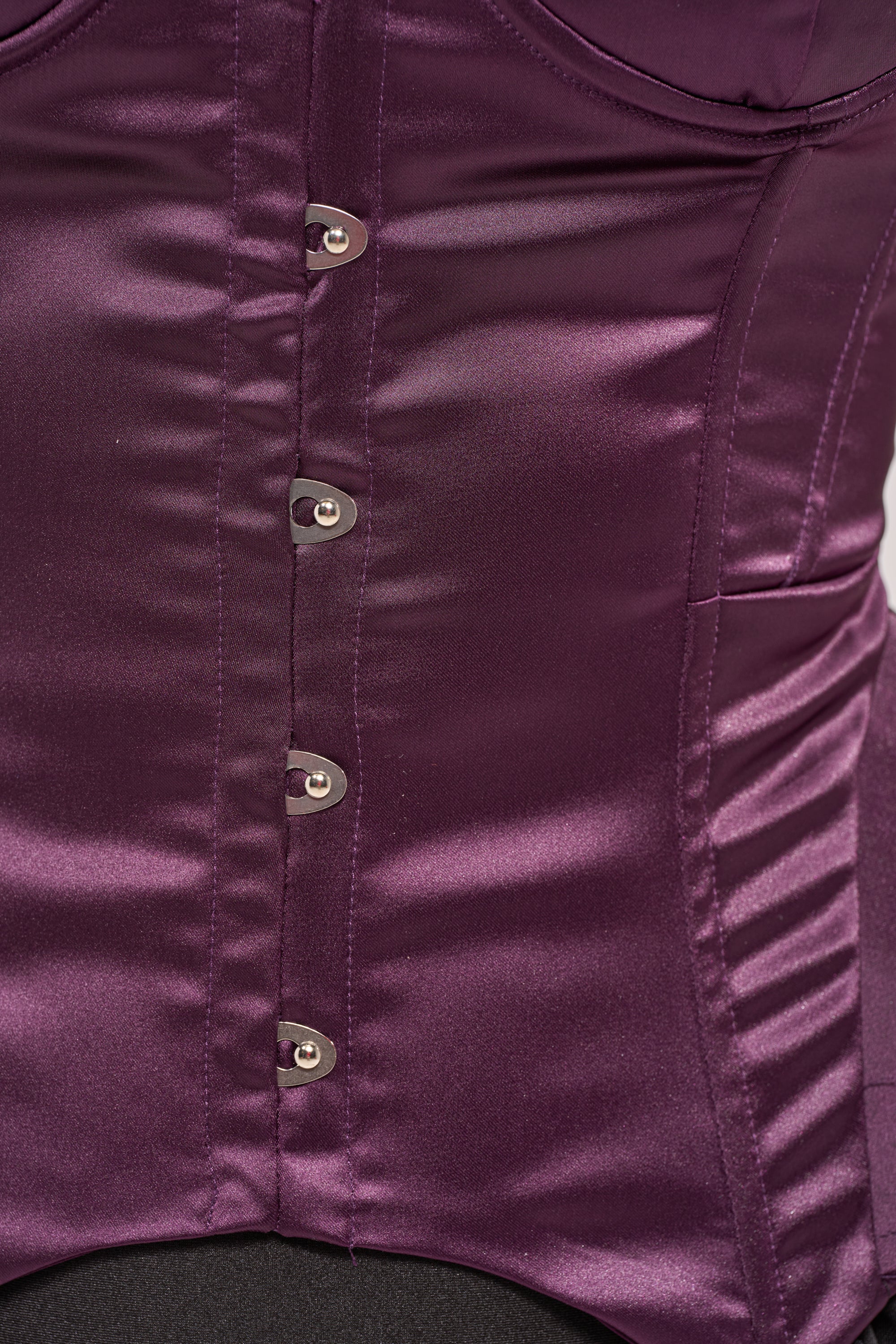 Close-up of the FLY ME OUT SATIN CORSET in shiny deep purple satin, featuring a peplum design, silver metal hook-and-eye front closures, and visible seam detailing. 5