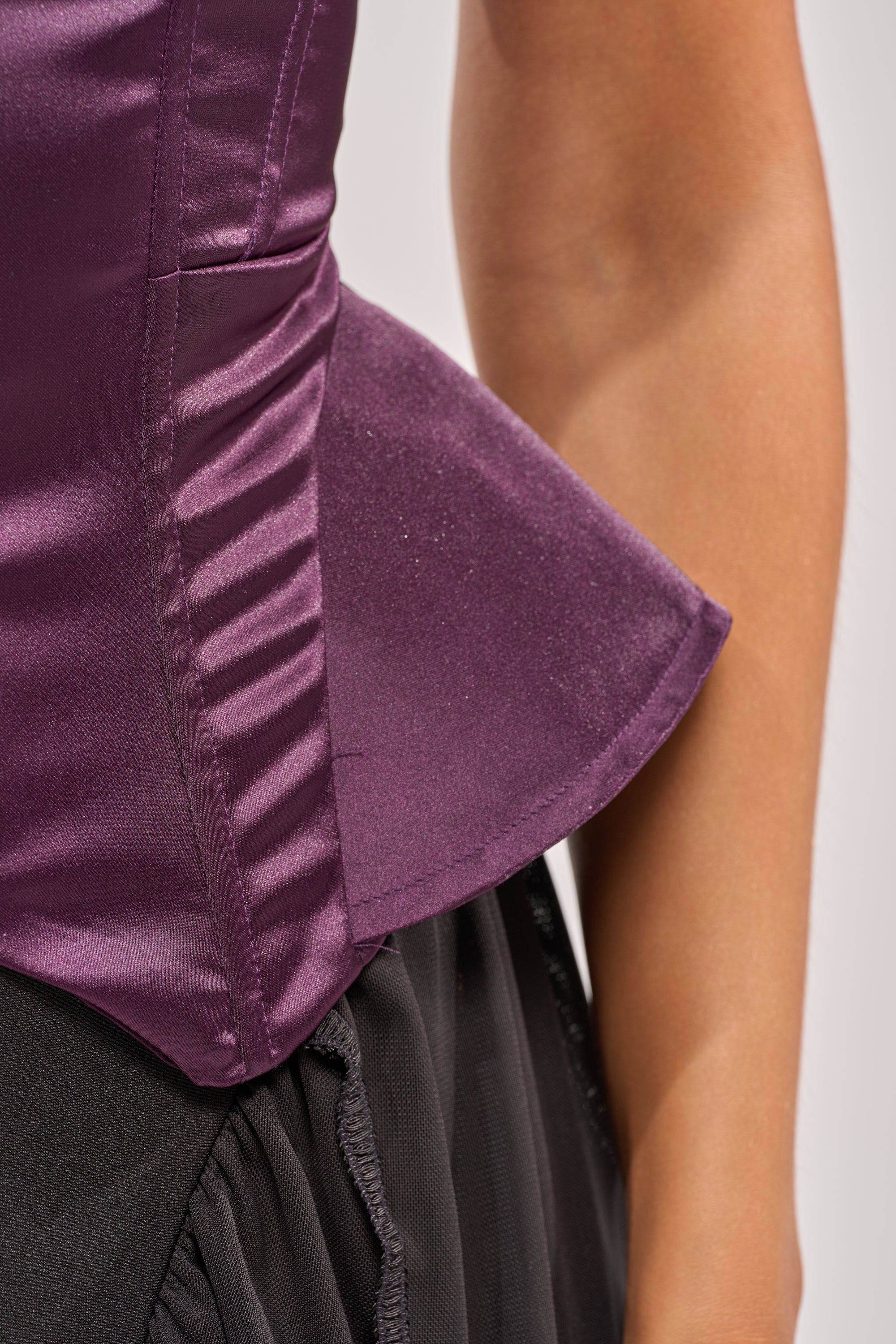 A close-up of a person wearing the FLY ME OUT SATIN CORSET in shiny purple with a black skirt, showing part of their bare arm against a neutral background. 6