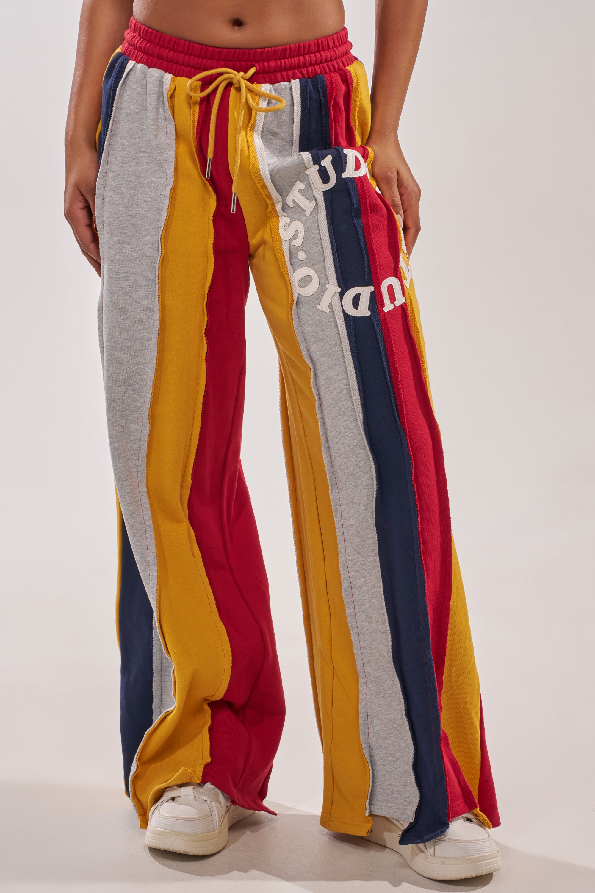 Someone stands against a plain background wearing STUDIO LIVING JOGGER sweatpants with wide legs, vertical red, yellow, gray, and navy stripes, "STUDIO" text on one leg, and white sneakers. 1