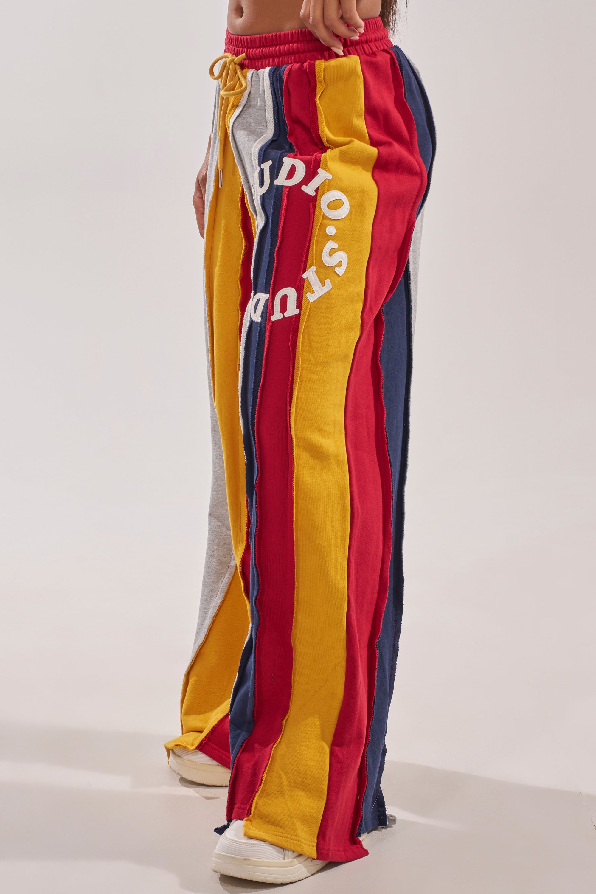 A person wears STUDIO LIVING JOGGER lounge pants featuring loose, colorful red, yellow, blue, and gray stripes with "STUDIOS" in white on the left leg, paired with white sneakers. 2