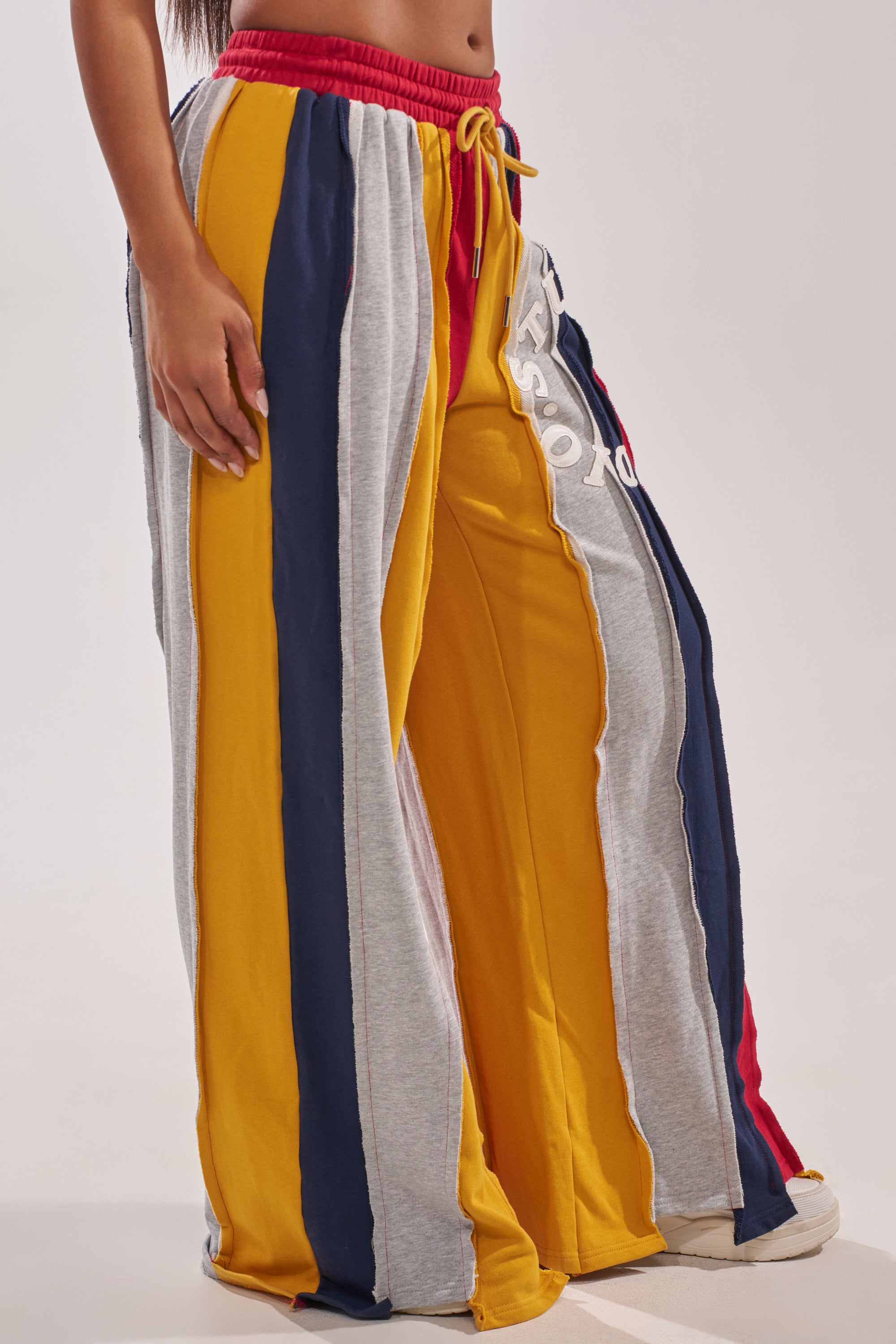 A person wearing the STUDIO LIVING JOGGER, wide-leg sweatpants with bold yellow, navy blue, gray, and red vertical stripes, an elastic red waistband, and white lettering on the left leg. Only the lower body is visible. 3