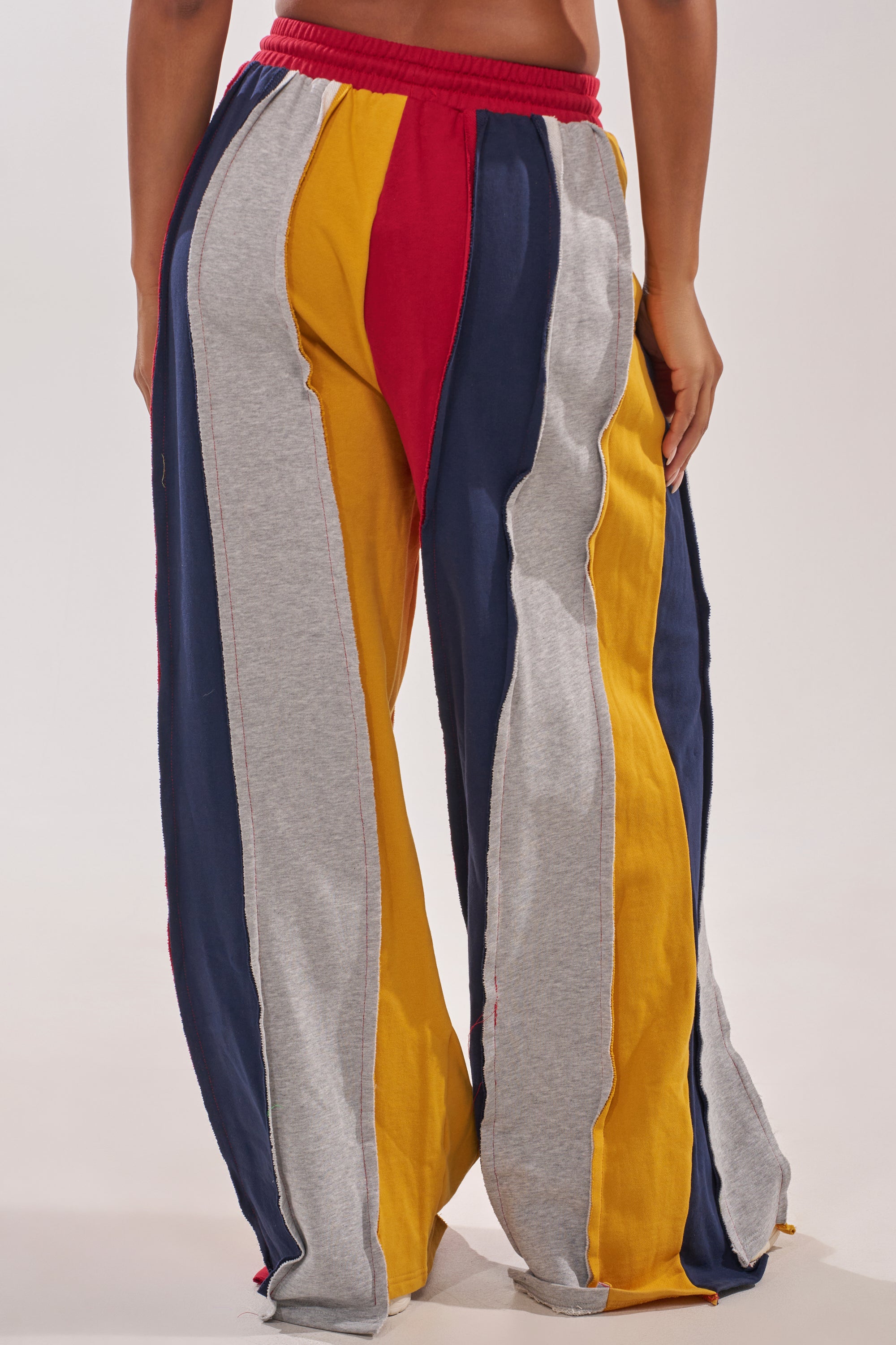 Model in STUDIO LIVING JOGGER: wide-leg, high-waisted lounge pants with bold color-block vertical stripes—red, yellow, navy blue, gray—elastic waistband and exposed seams for a relaxed fit. 4