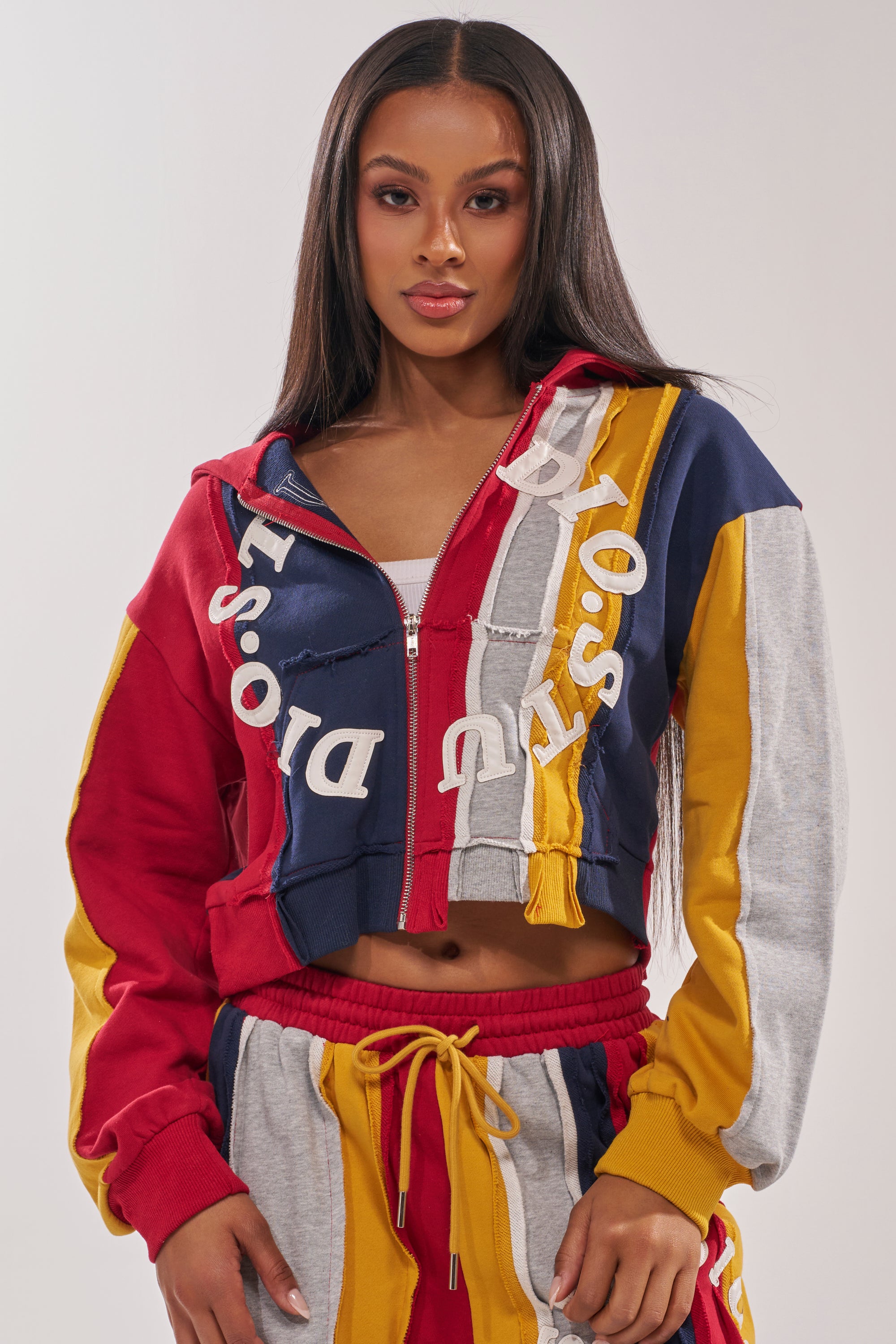 A woman with long straight hair confidently models the STUDIO LIVING SWEATSHIRT, a color-blocked zip-up hoodie and matching sweatpants in red, navy, yellow, and gray with bold white lettering, against a plain light background. 0