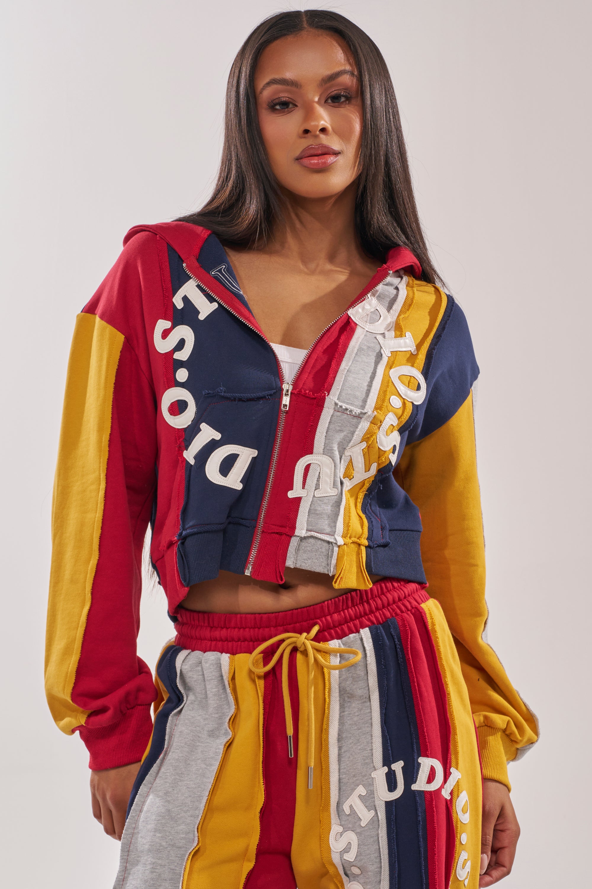 A woman wears the STUDIO LIVING SWEATSHIRT, a color-block hoodie and sweatpants set featuring red, yellow, navy, and gray panels with bold white "DISO" lettering. She has long straight hair and a neutral expression. 1