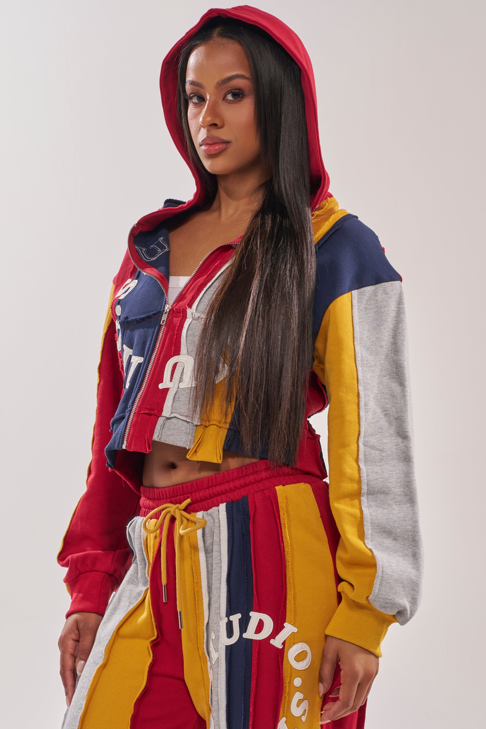 A woman stands facing forward in the STUDIO LIVING SWEATSHIRT, a bold, colorful patchwork hoodie with matching sweatpants in red, yellow, blue, and gray. She has the hood up and her long straight hair draped over one shoulder. 3