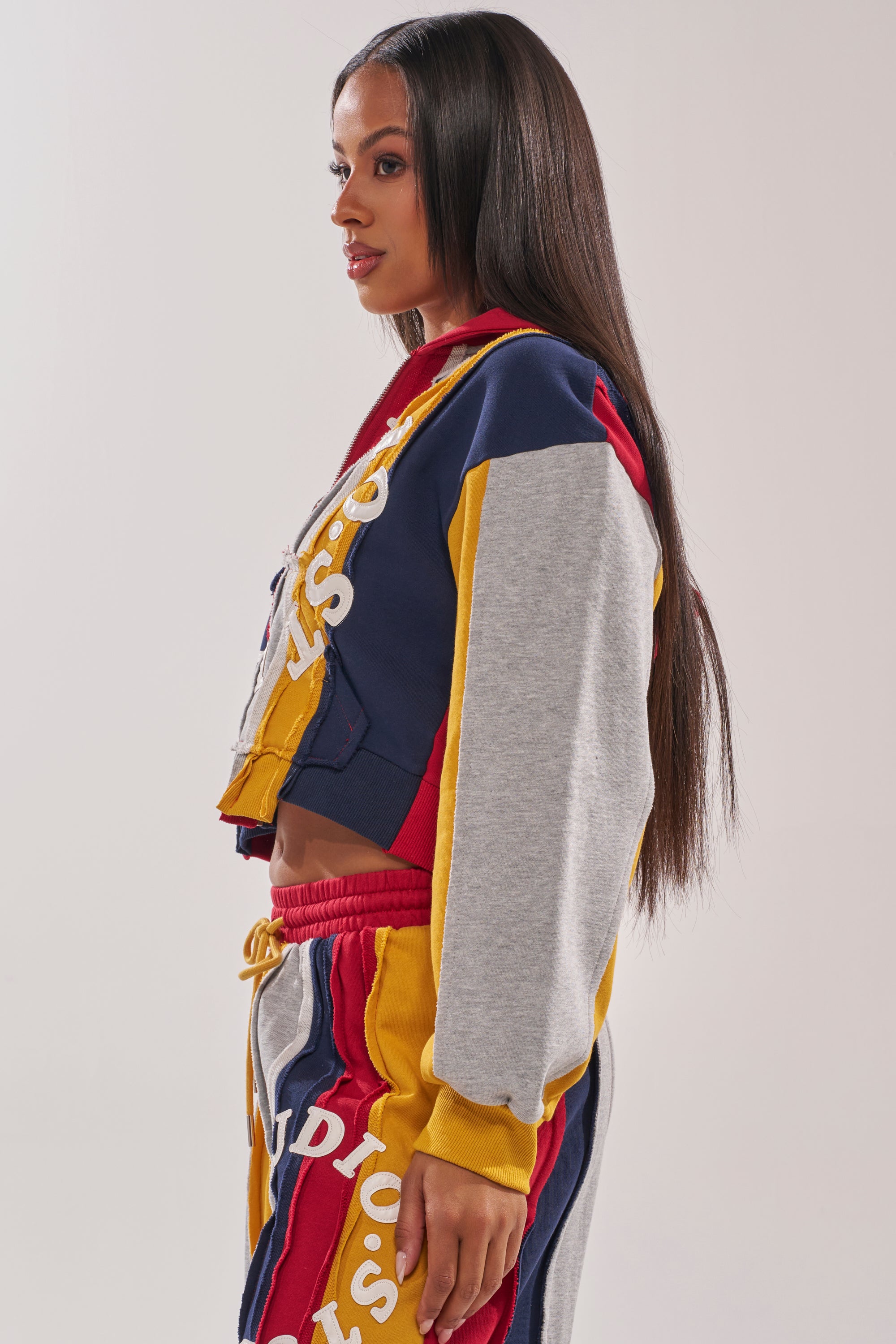 A woman with long, straight hair models the STUDIO LIVING SWEATSHIRT—a colorful tracksuit top with navy, red, yellow, and gray panels and bold white lettering—posing in profile against a plain light background. 4
