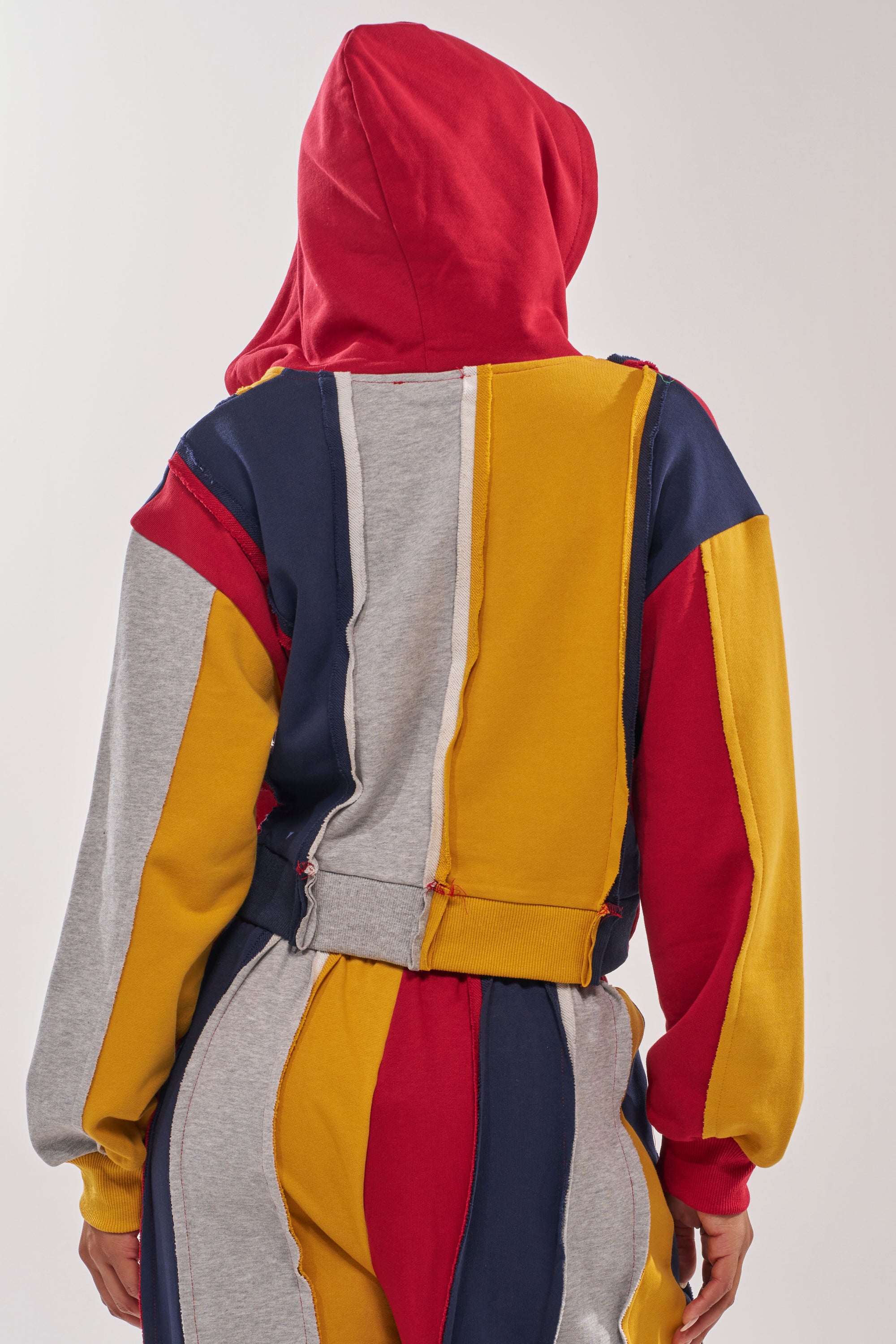 A person wears the STUDIO LIVING SWEATSHIRT, featuring red, yellow, gray, and navy color-blocked panels; hood up and back to the camera, highlighting exposed seams and a casual streetwear look. 5