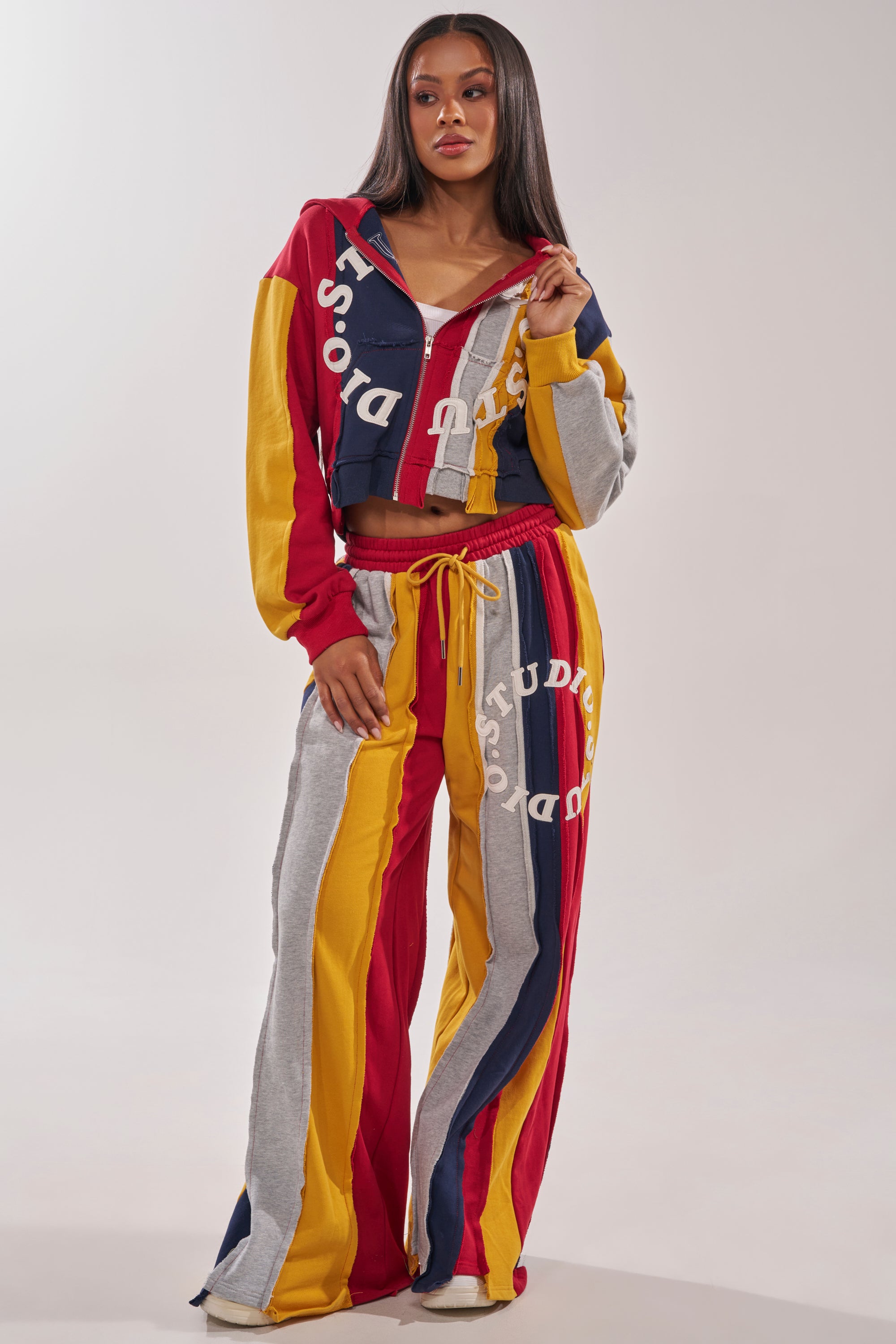 A woman in trendy streetwear wears the STUDIO LIVING SWEATSHIRT, a colorful color-blocked hoodie with red, yellow, gray, and navy blue stripes and white “Sore Studio” text, paired with wide-leg pants against a plain light background. 6