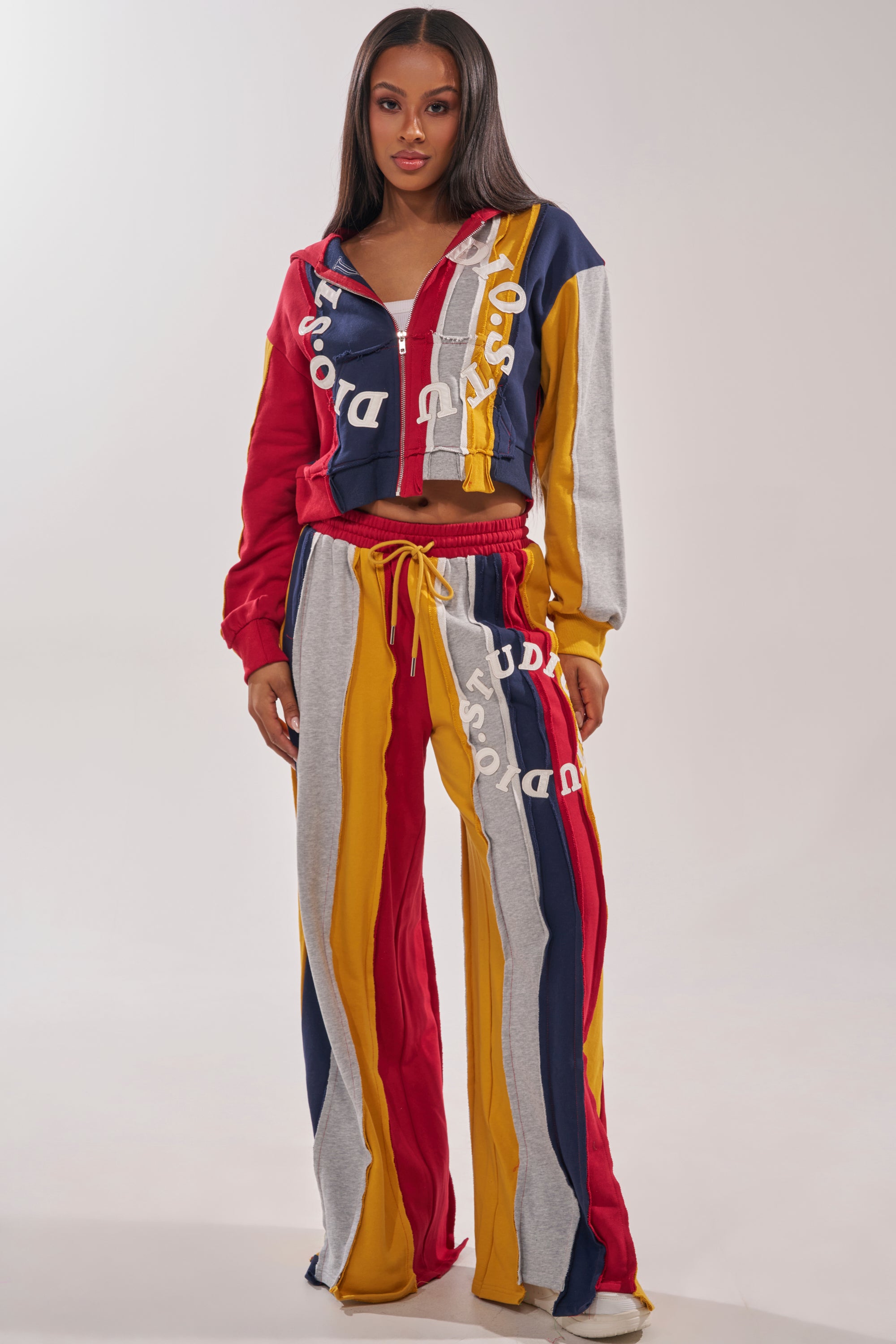 A woman wears the STUDIO LIVING SWEATSHIRT, a vibrant, streetwear-inspired color-block hoodie in red, yellow, navy blue, and gray stripes with matching drawstring pants and bold white text accents. 7