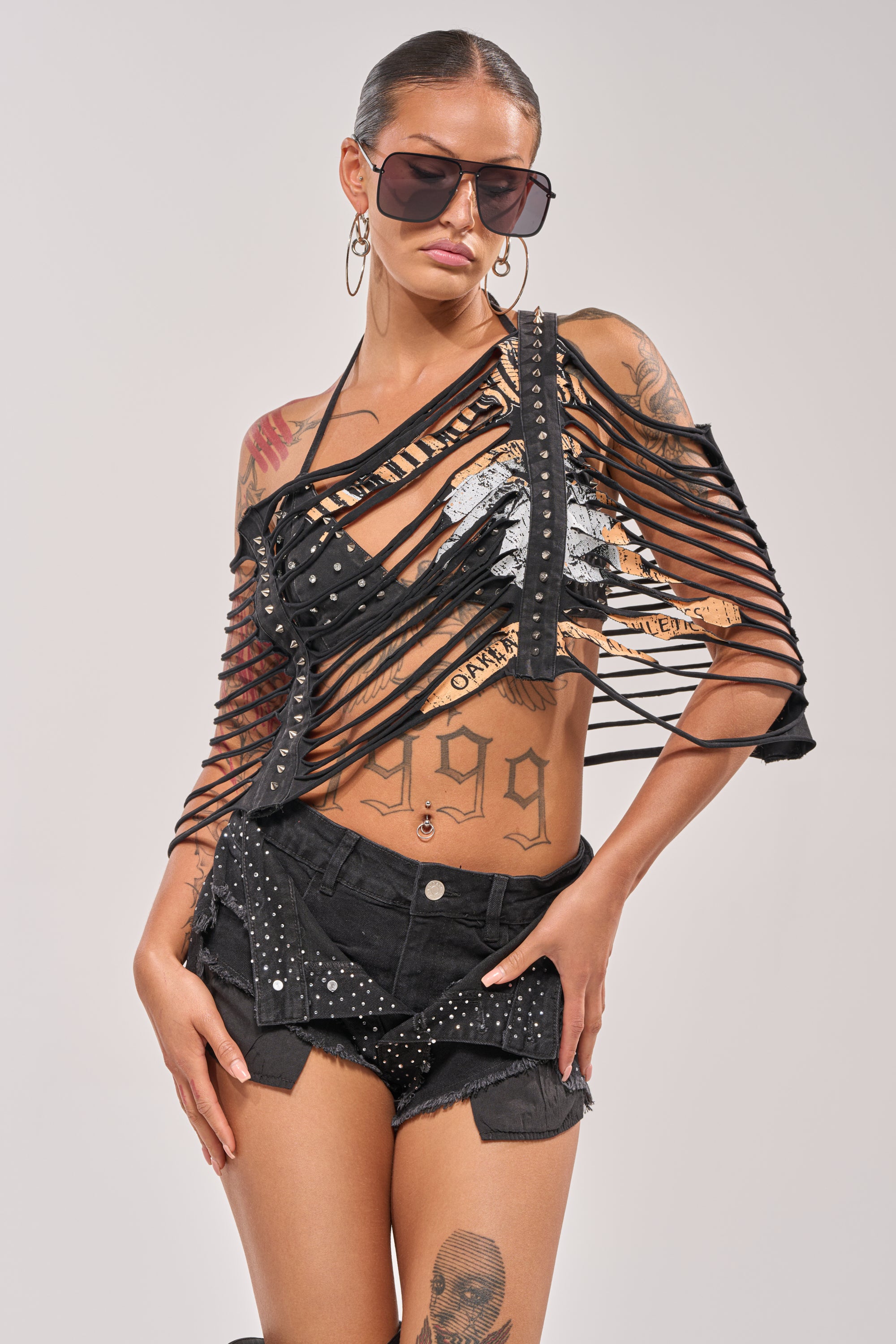 A woman with festival styling wears large sunglasses, hoop earrings, studded black shorts, and the CHELLA DISTRESSED TOP—a punk-inspired ripped black top with graphic details. Her slicked-back hair and tattoos complete the bold look. 2