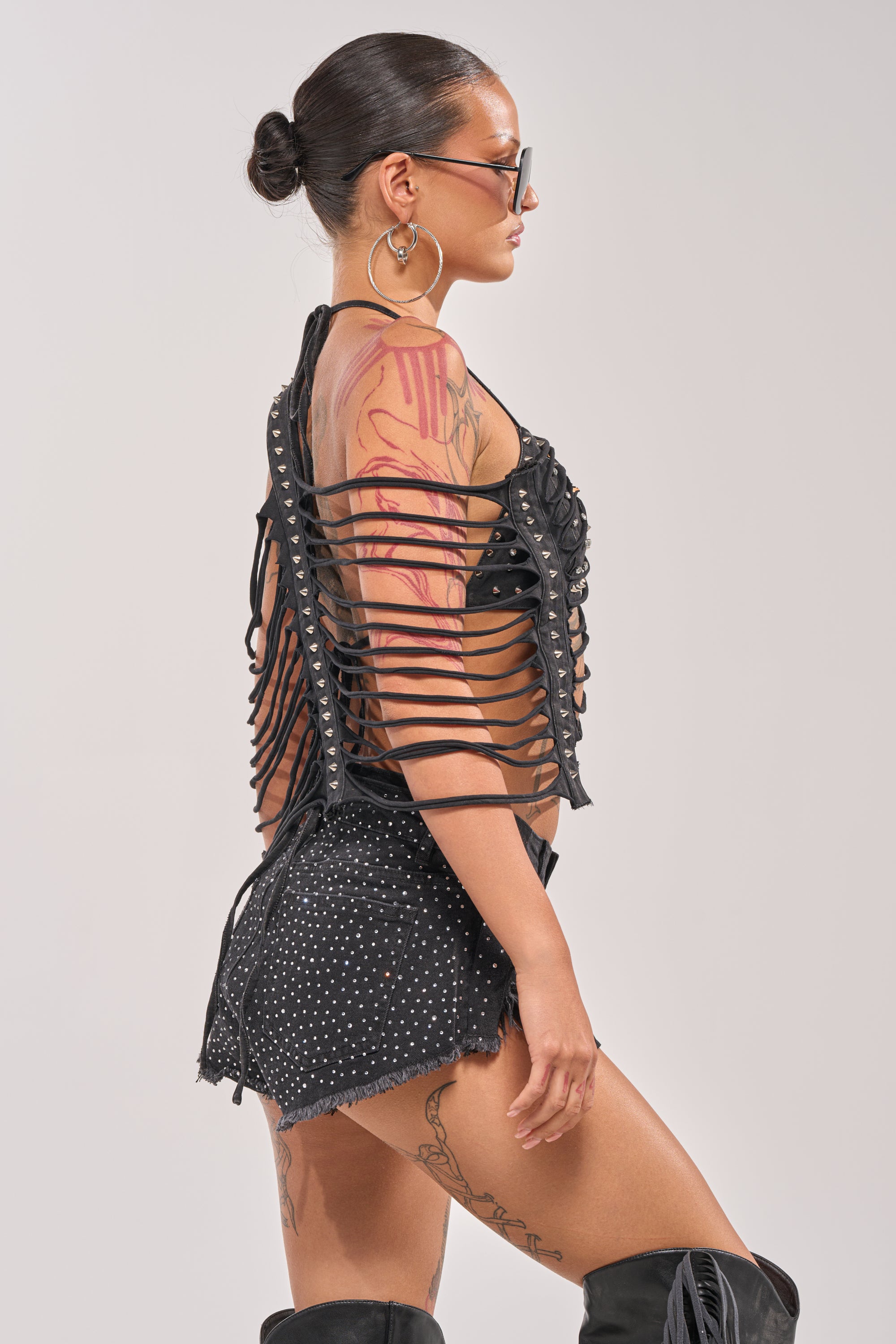 A woman with sunglasses and hoop earrings poses sideways in a punk-inspired look, featuring the CHELLA DISTRESSED TOP, studded shorts, knee-high boots, visible tattoos on her arm and leg, and her hair styled in a bun for bold festival flair. 4