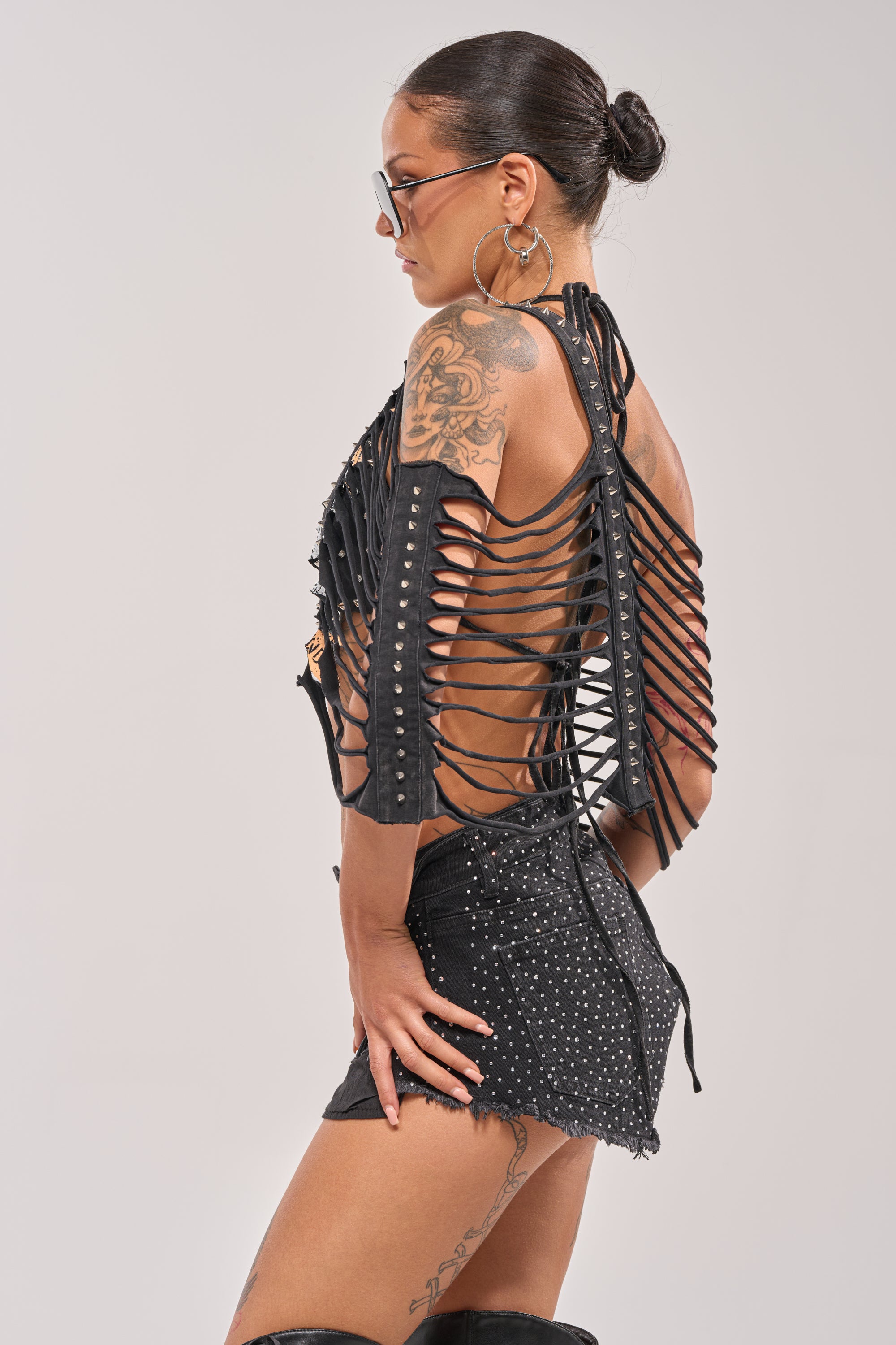 Woman with tattoos and large hoop earrings poses in profile, wearing the CHELLA DISTRESSED TOP with grommets, black studded shorts, and sunglasses. Her hair is in a sleek bun against a plain background for a cool punk-inspired vibe. 5