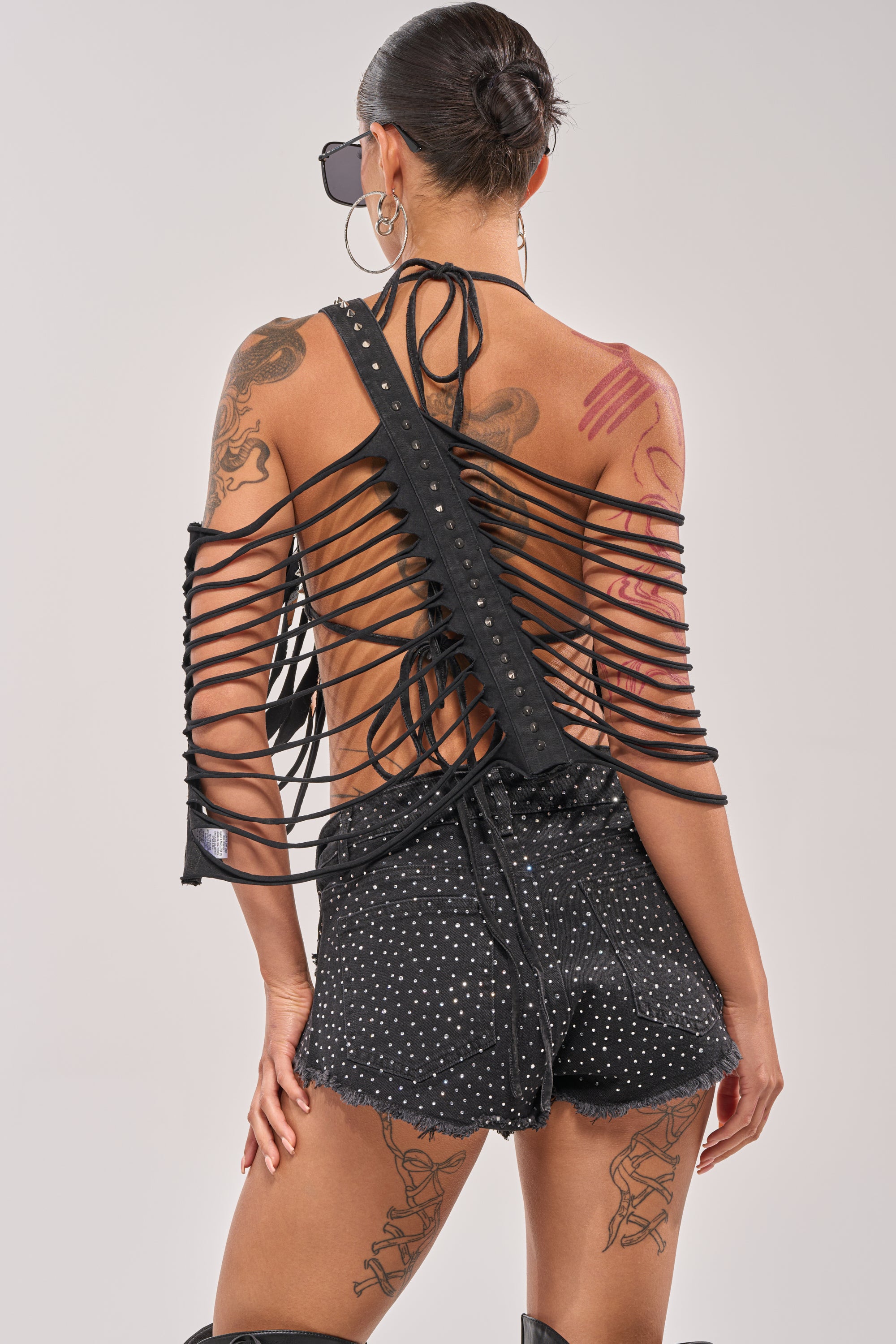 Wearing the CHELLA DISTRESSED TOP, a tattooed person pairs it with black sunglasses and black studded polka dot shorts, facing away from the camera against a plain background. 6
