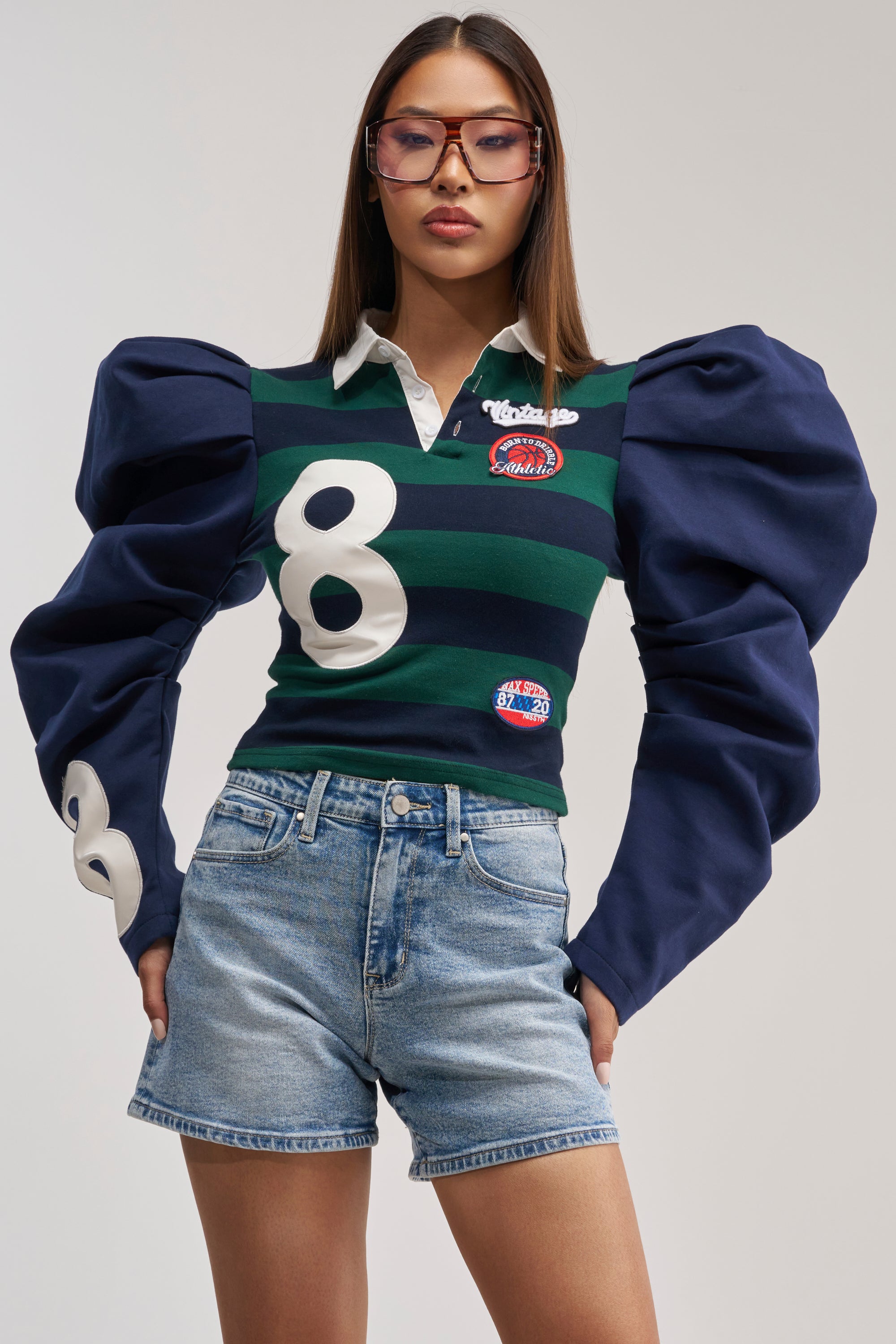 A woman wearing glasses and the COLLEGE DAYS STRIPE POLO—with exaggerated puff sleeves and varsity patches—paired with denim shorts, stands confidently with her hands on her hips against a plain background.