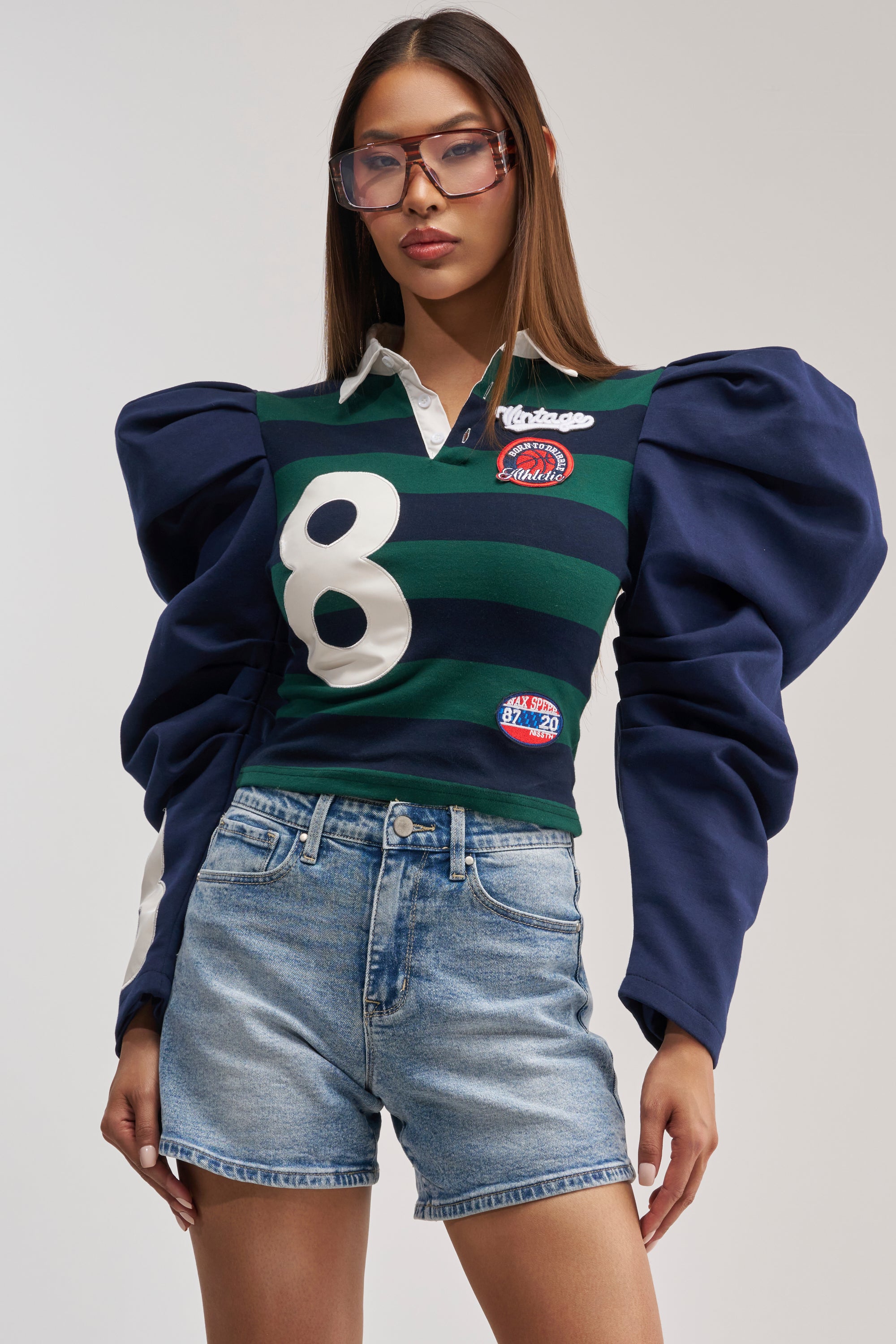 A woman stands against a plain background wearing oversized glasses, light blue denim shorts, and the COLLEGE DAYS STRIPE POLO—an oversized green and navy striped puff sleeve top with a white number 8 varsity patch.